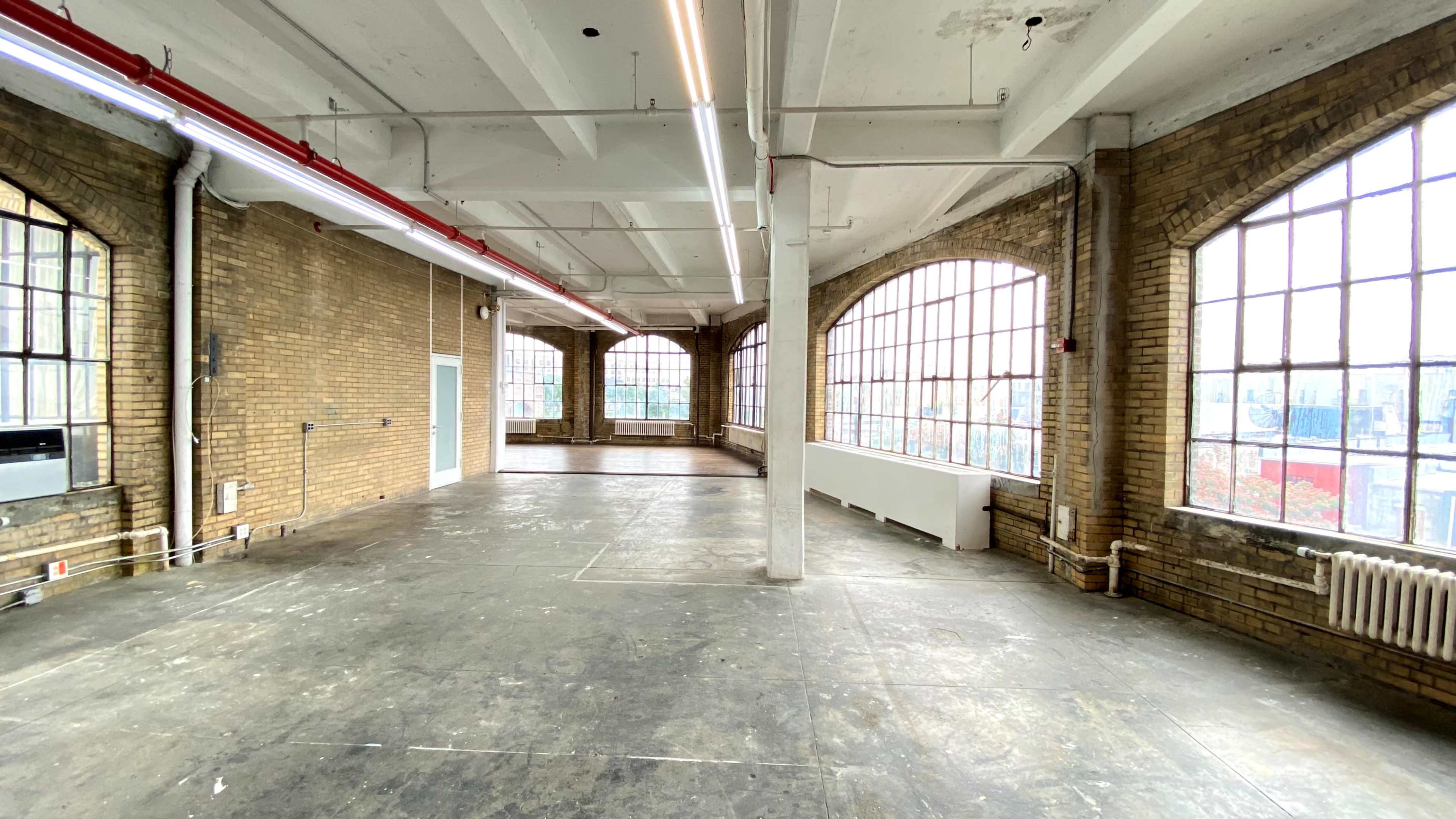 A spacious, empty room with large windows, exposed brick walls, and concrete flooring.