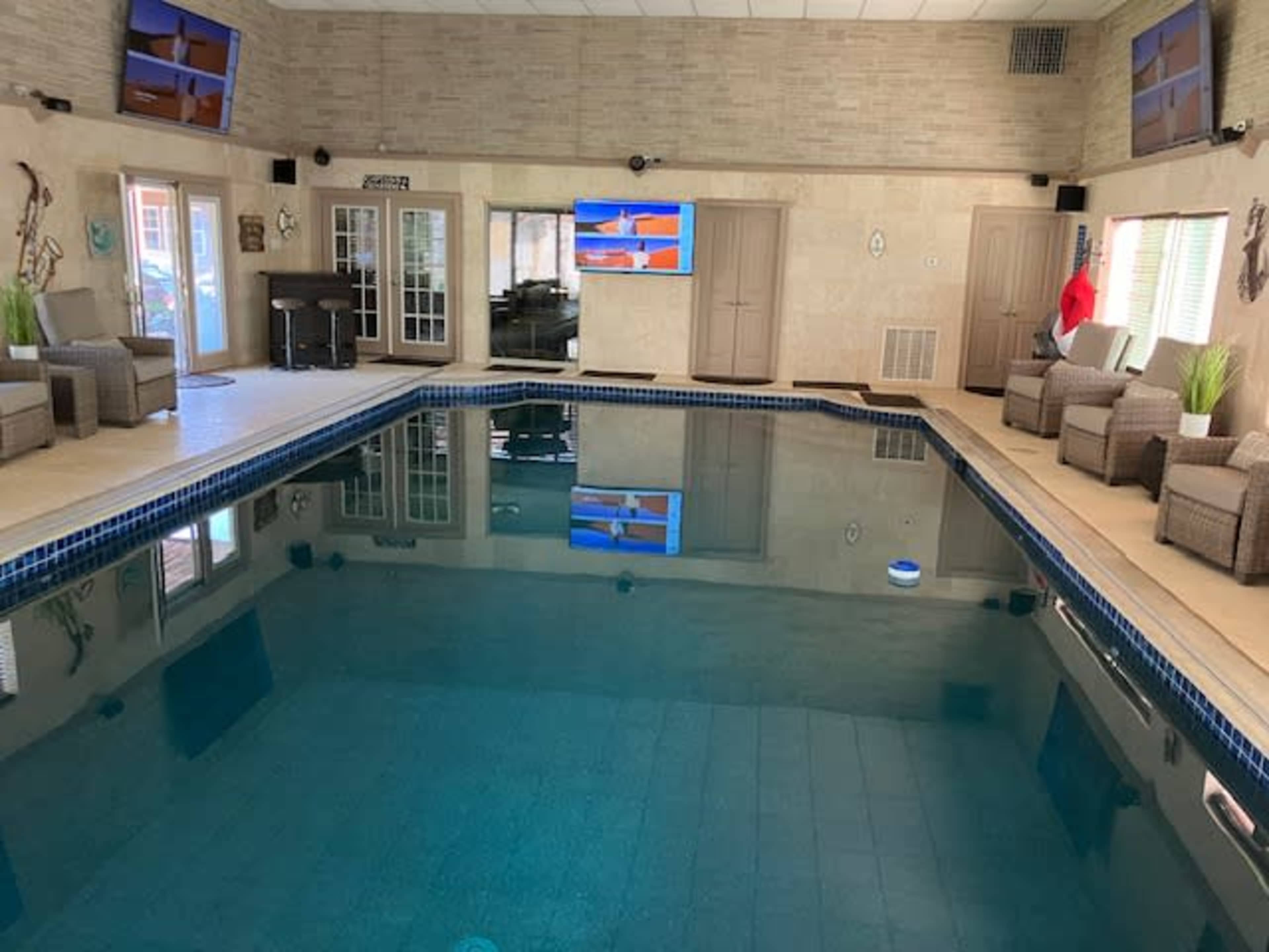The image shows an indoor swimming pool area with lounge chairs, large windows, and flat-screen televisions mounted on the walls.