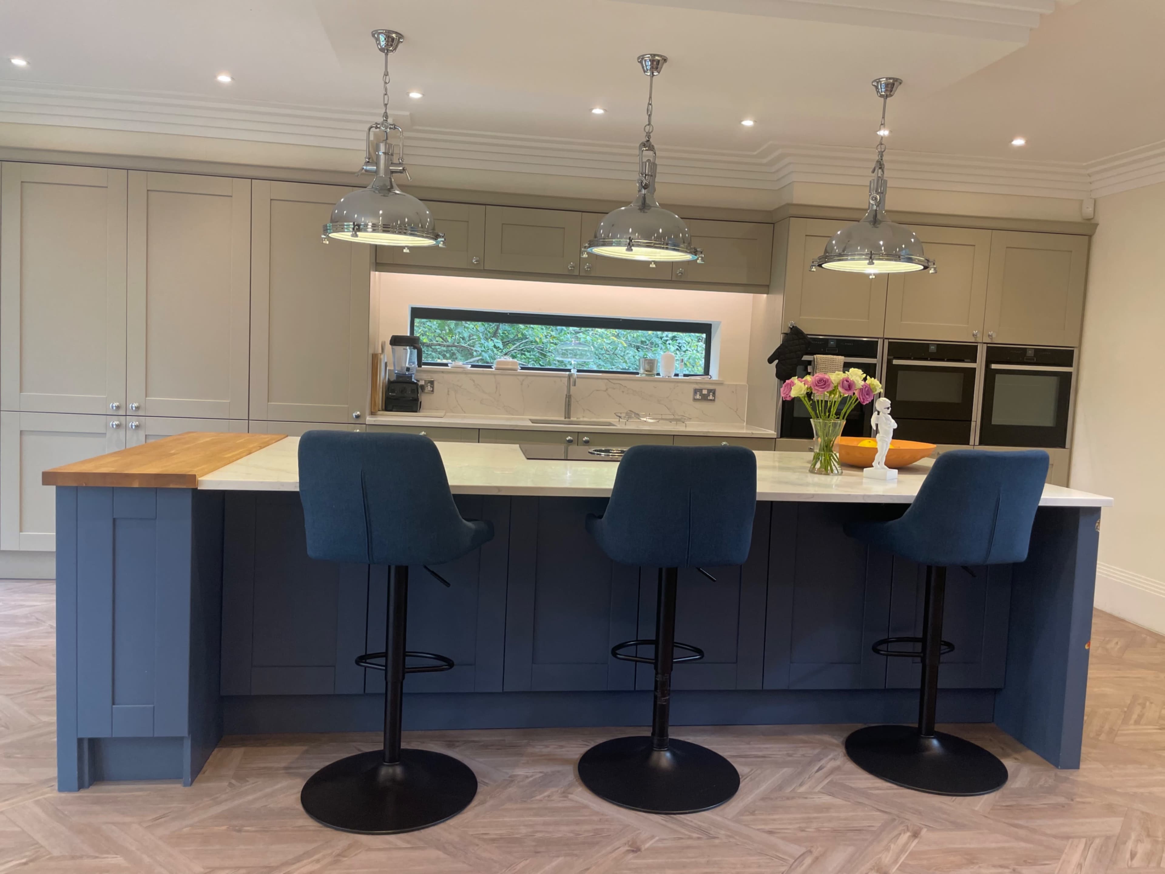 The image shows a modern kitchen featuring a central island with three tall blue bar stools, pendant lights above, and a mix of cabinetry in light and dark colors.