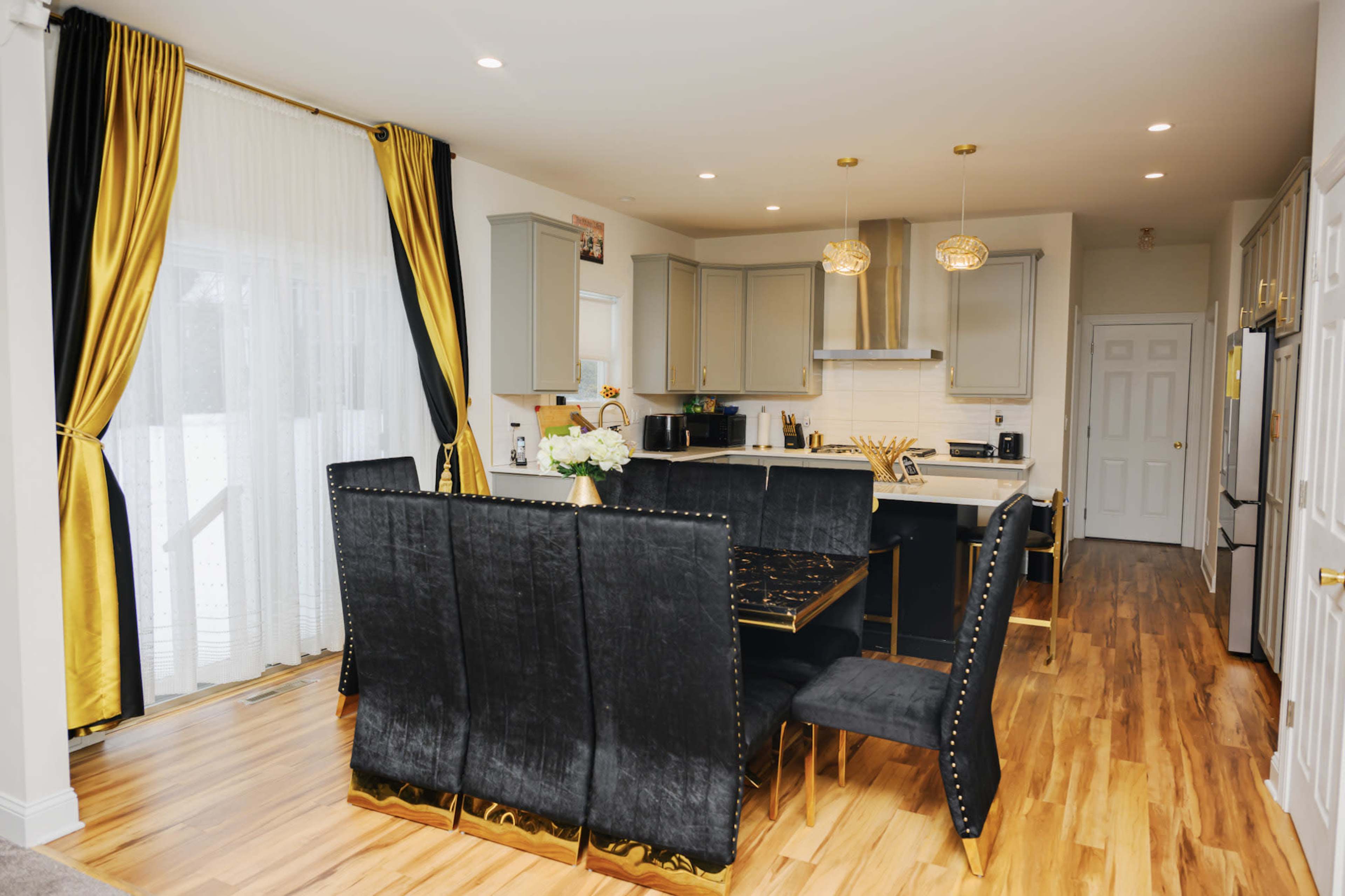 The image shows a modern kitchen and dining area featuring a gray and gold color scheme, with a large black dining table surrounded by upholstered chairs and a bright window in the background.
