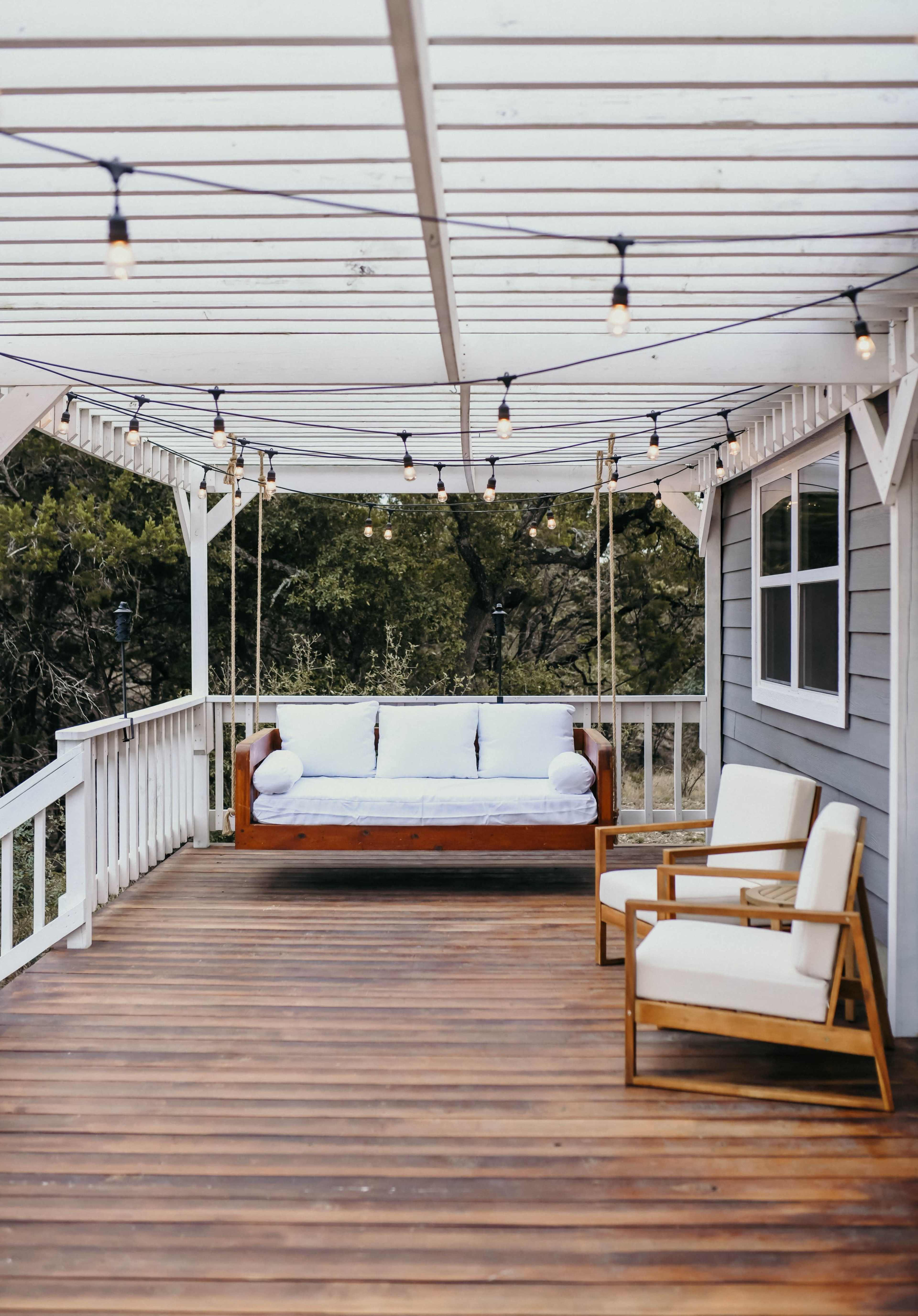 A covered wooden deck with a swing bench and two white cushioned chairs, adorned with string lights overhead.
