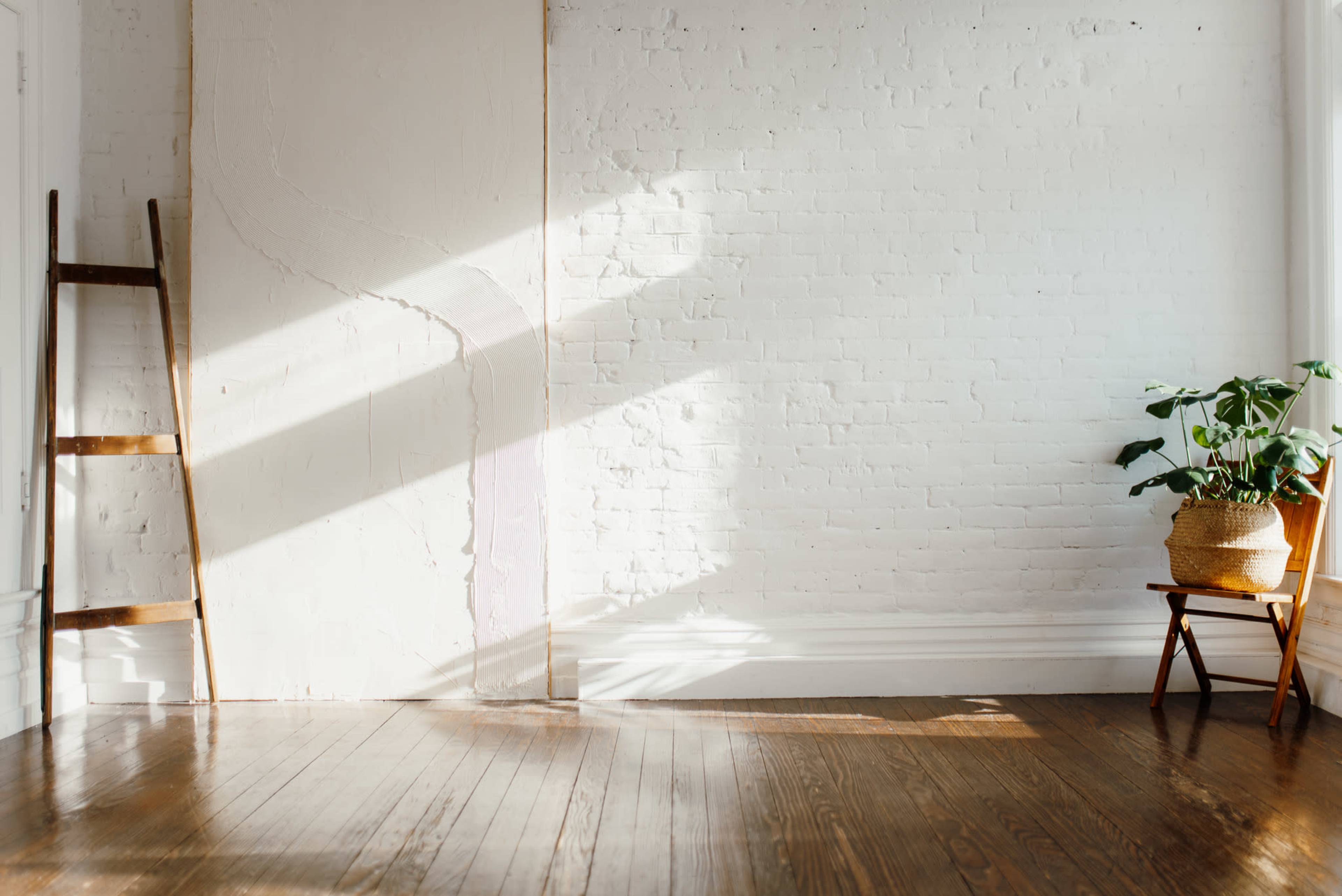 A sunlit room features a wooden ladder against a white brick wall and a potted plant next to a chair on a polished wooden floor.