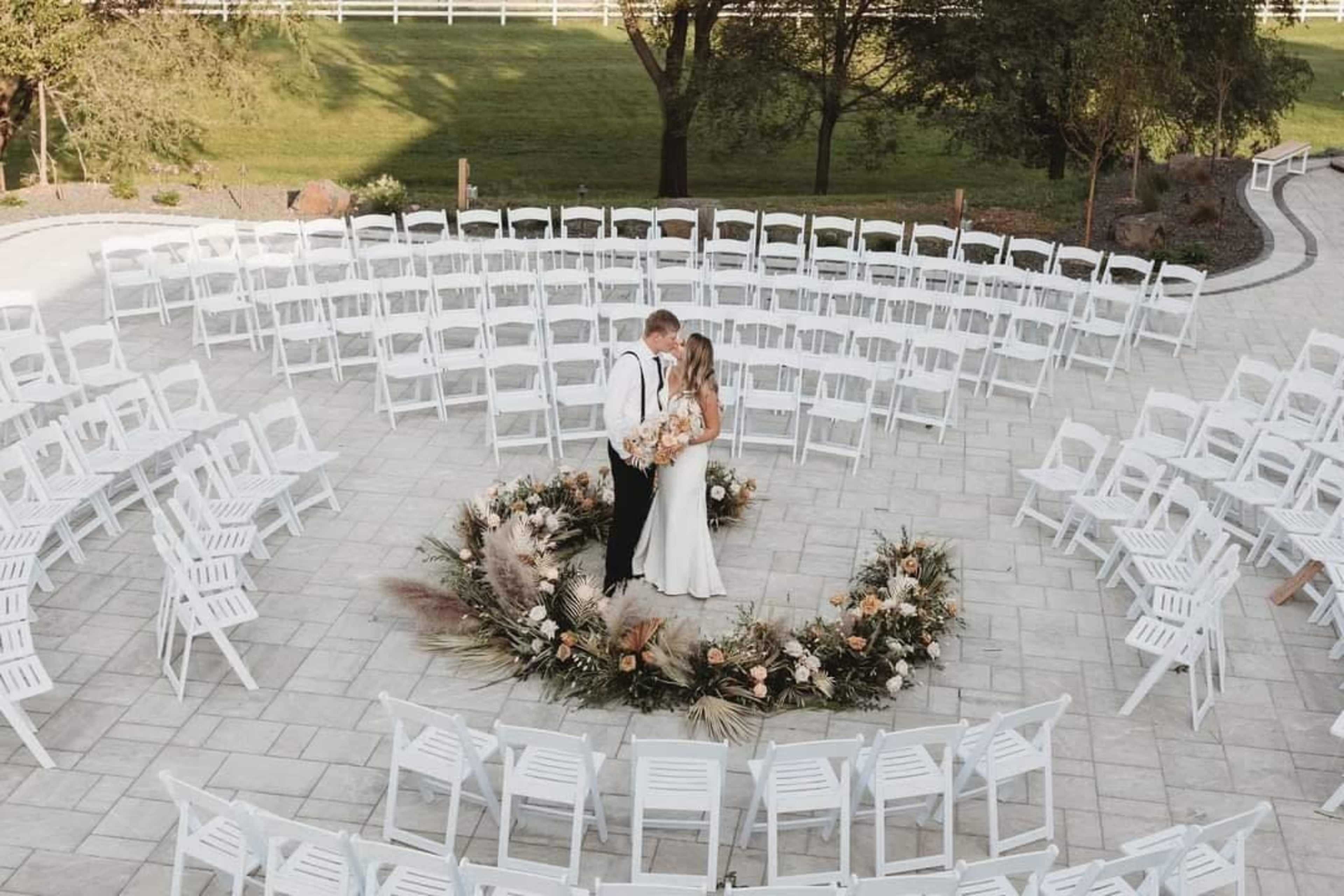 A couple stands together in the center of a circular arrangement of white chairs, surrounded by a floral decoration on a stone patio.