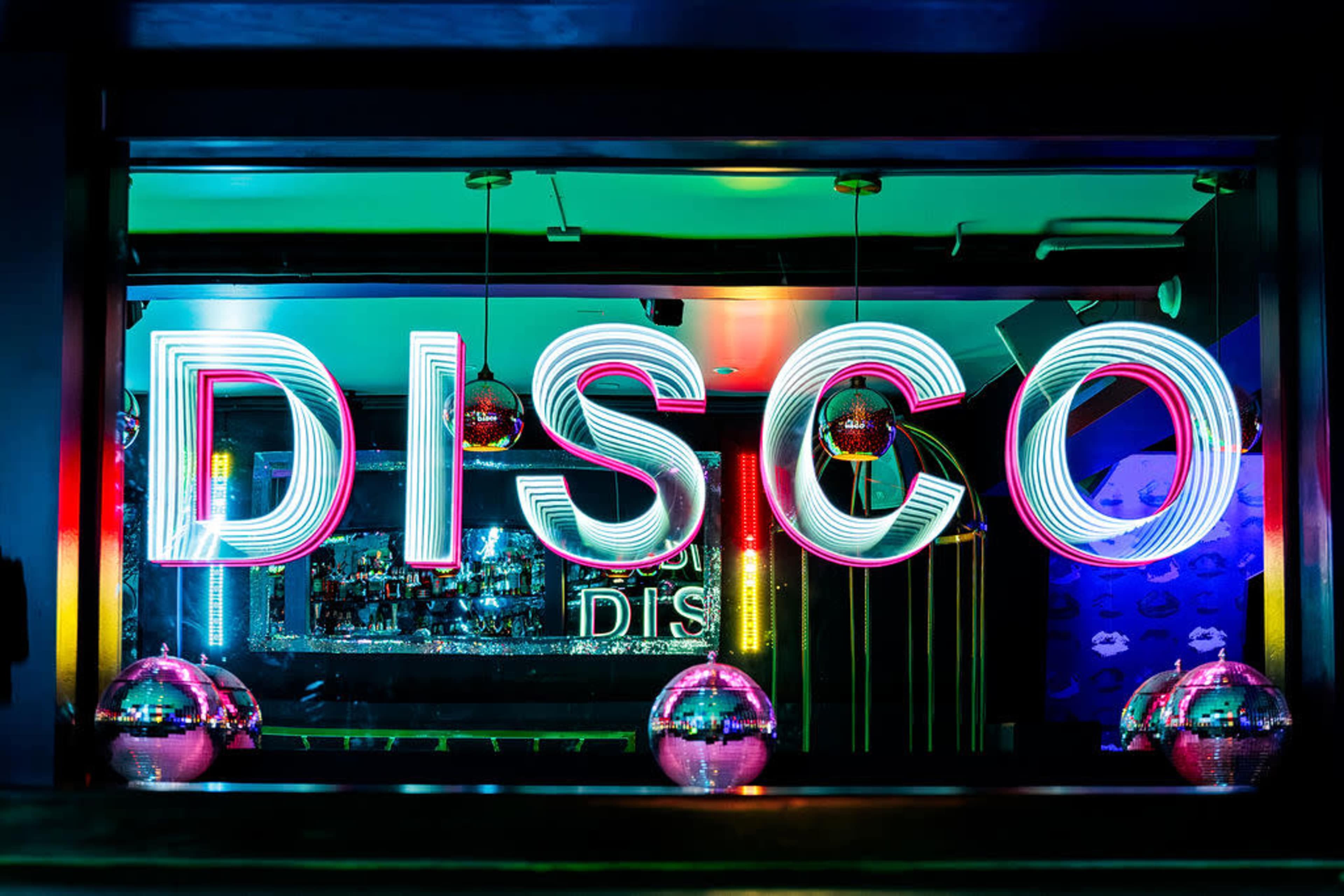 The image shows a brightly lit bar window with the word "DISCO" in neon lights, surrounded by colorful decorations and disco balls.