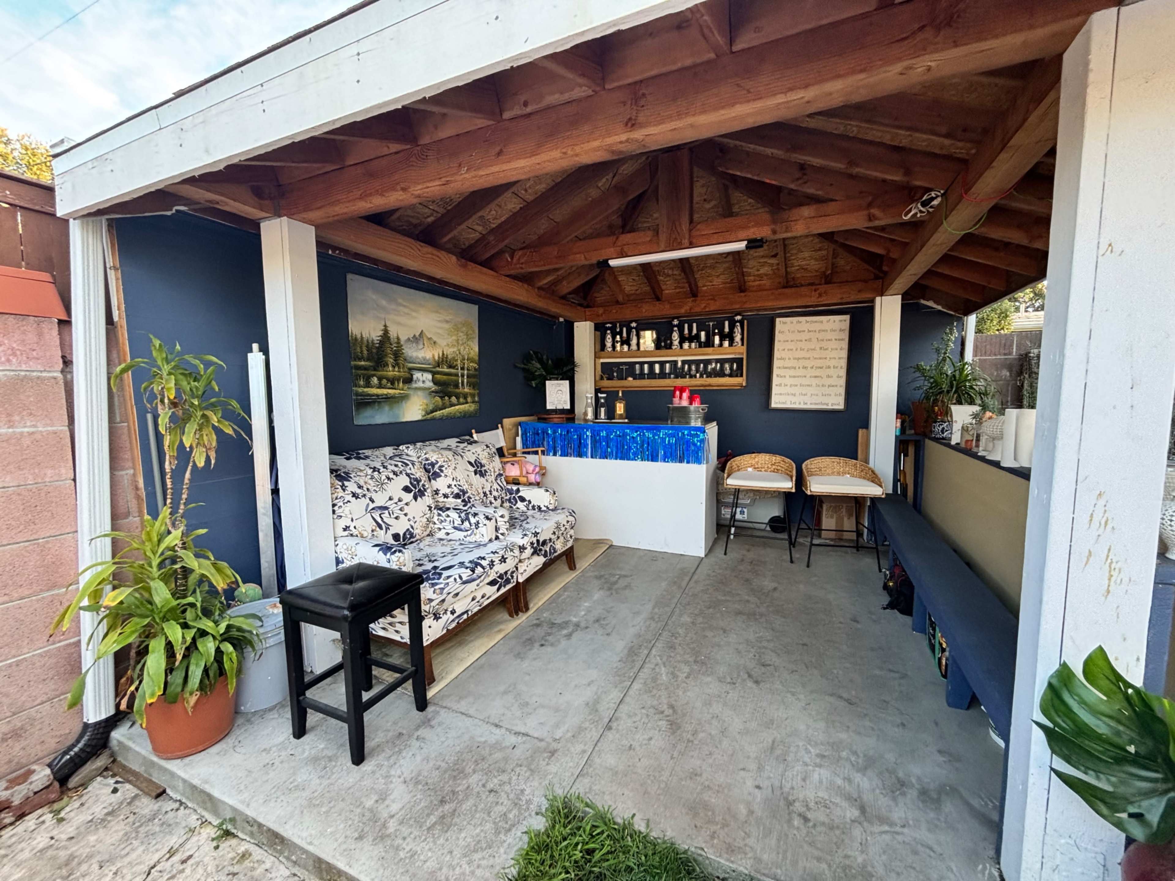 The image shows a small outdoor lounge area featuring a floral-patterned sofa, a bar with bottles against the wall, and a seating area under a wooden roof.