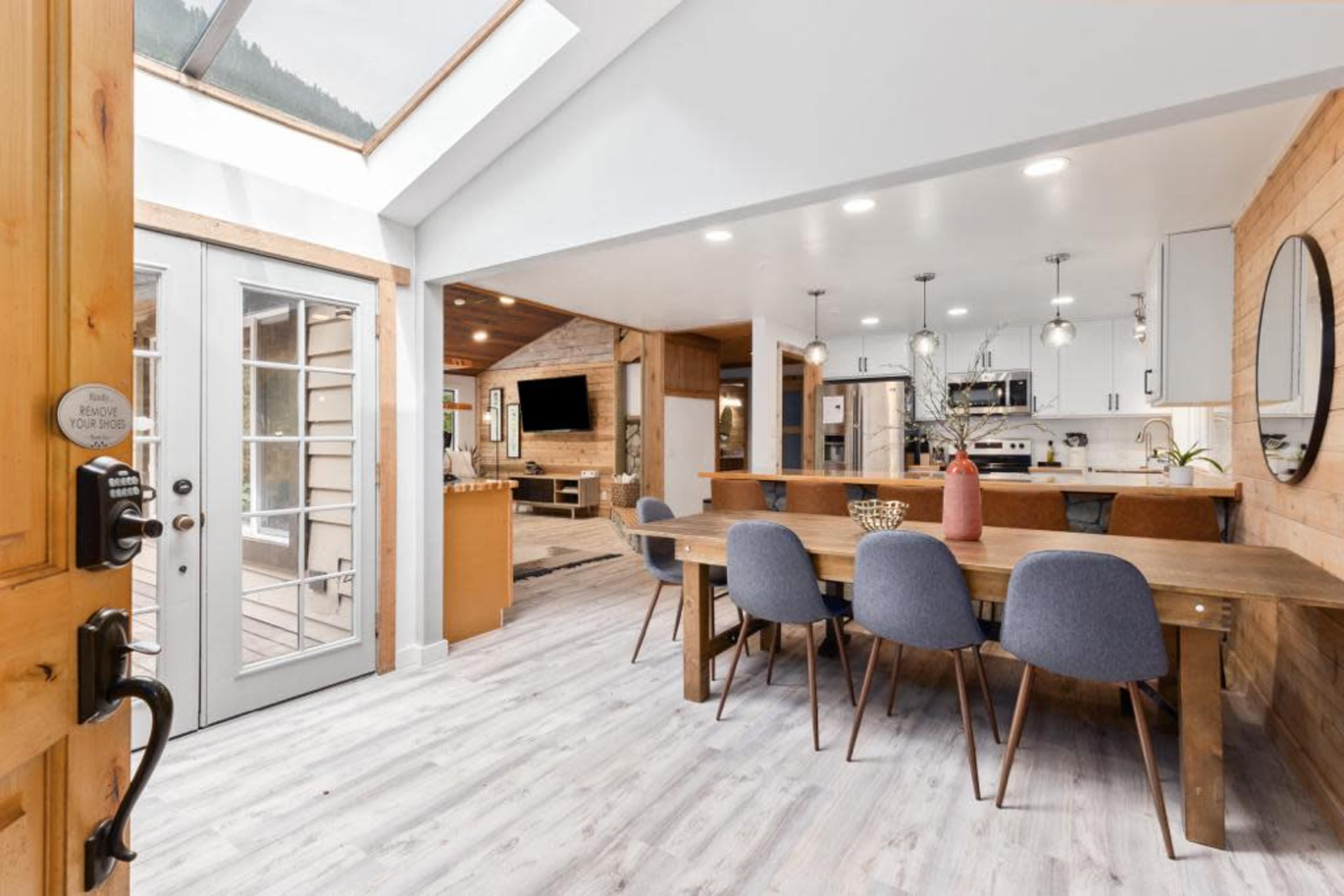 A modern kitchen and dining area featuring a long wooden table surrounded by gray chairs, with large windows that allow natural light to fill the space.