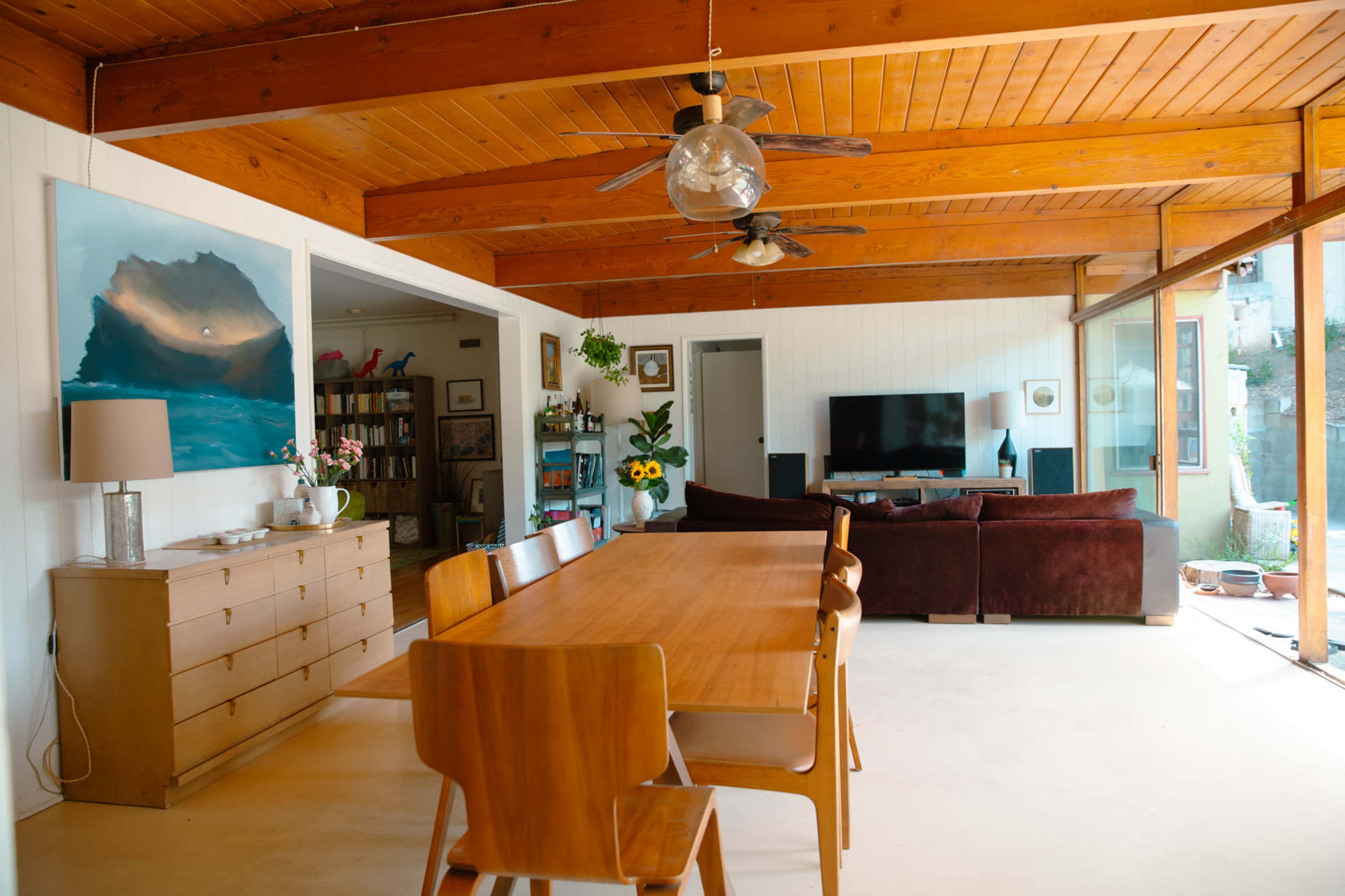 A dining area features a wooden table surrounded by chairs, with a living space visible in the background containing a couch, television, and artwork.