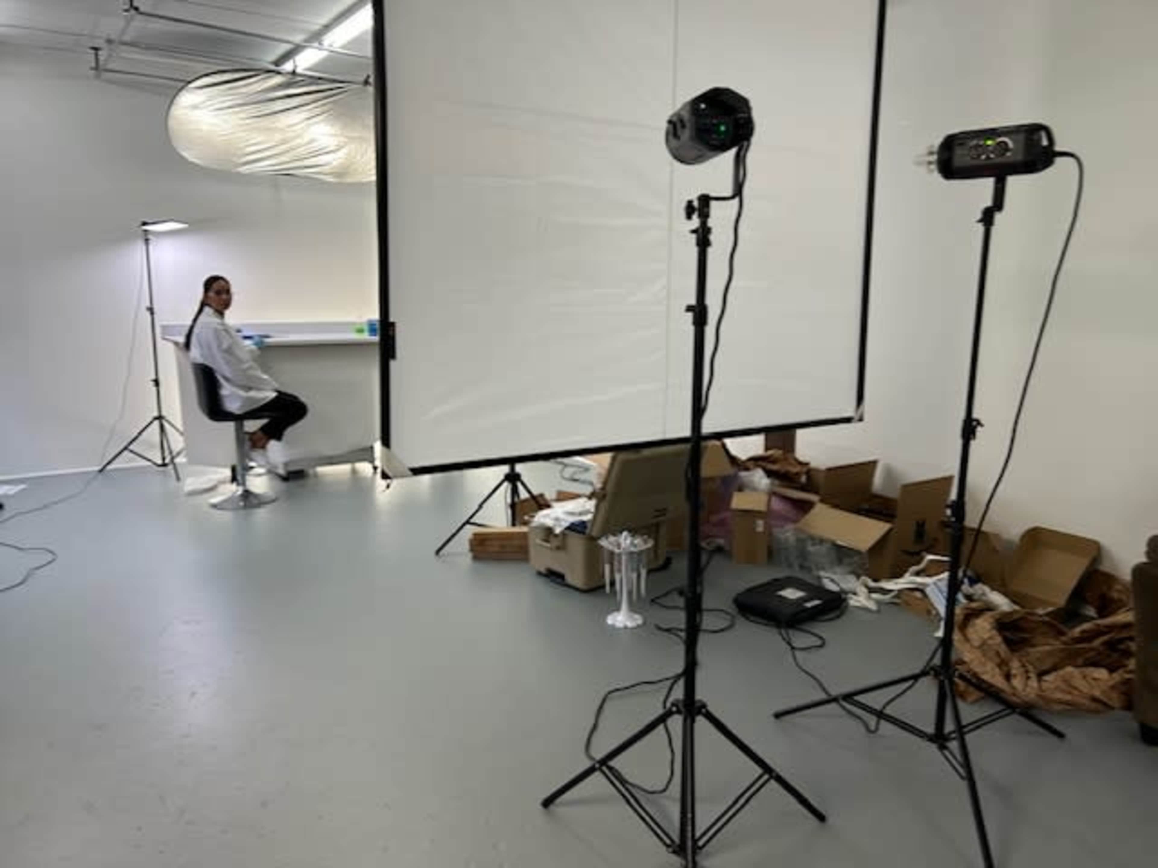 A person sits on a stool in a studio setting equipped with photography lighting and a backdrop.