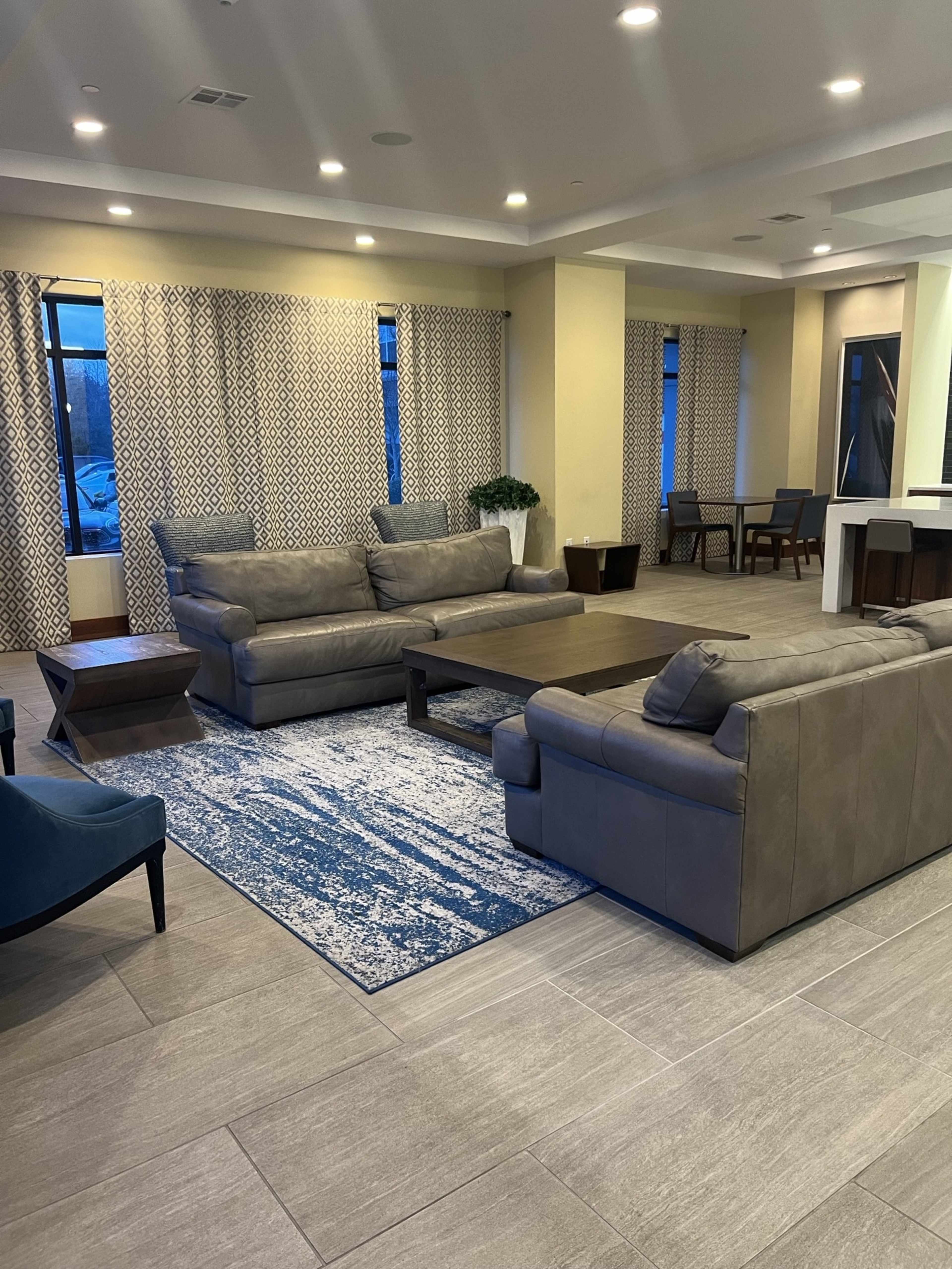 A modern lounge area featuring sectional sofas, a coffee table, a rug, and dining chairs, with large windows dressed in patterned curtains.