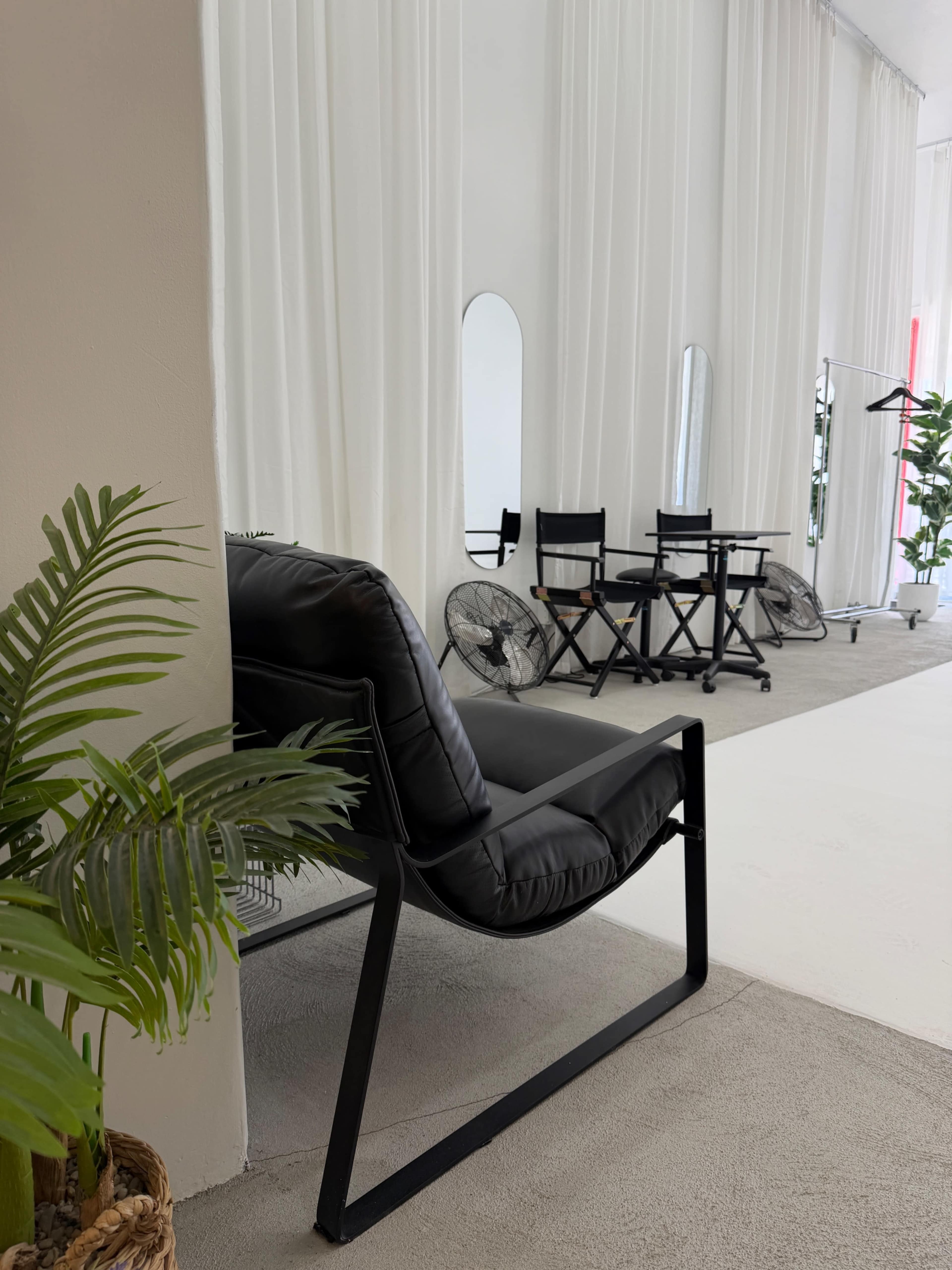 The image features a black lounge chair positioned next to a potted plant, with white curtains and a small table and chairs set in the background.