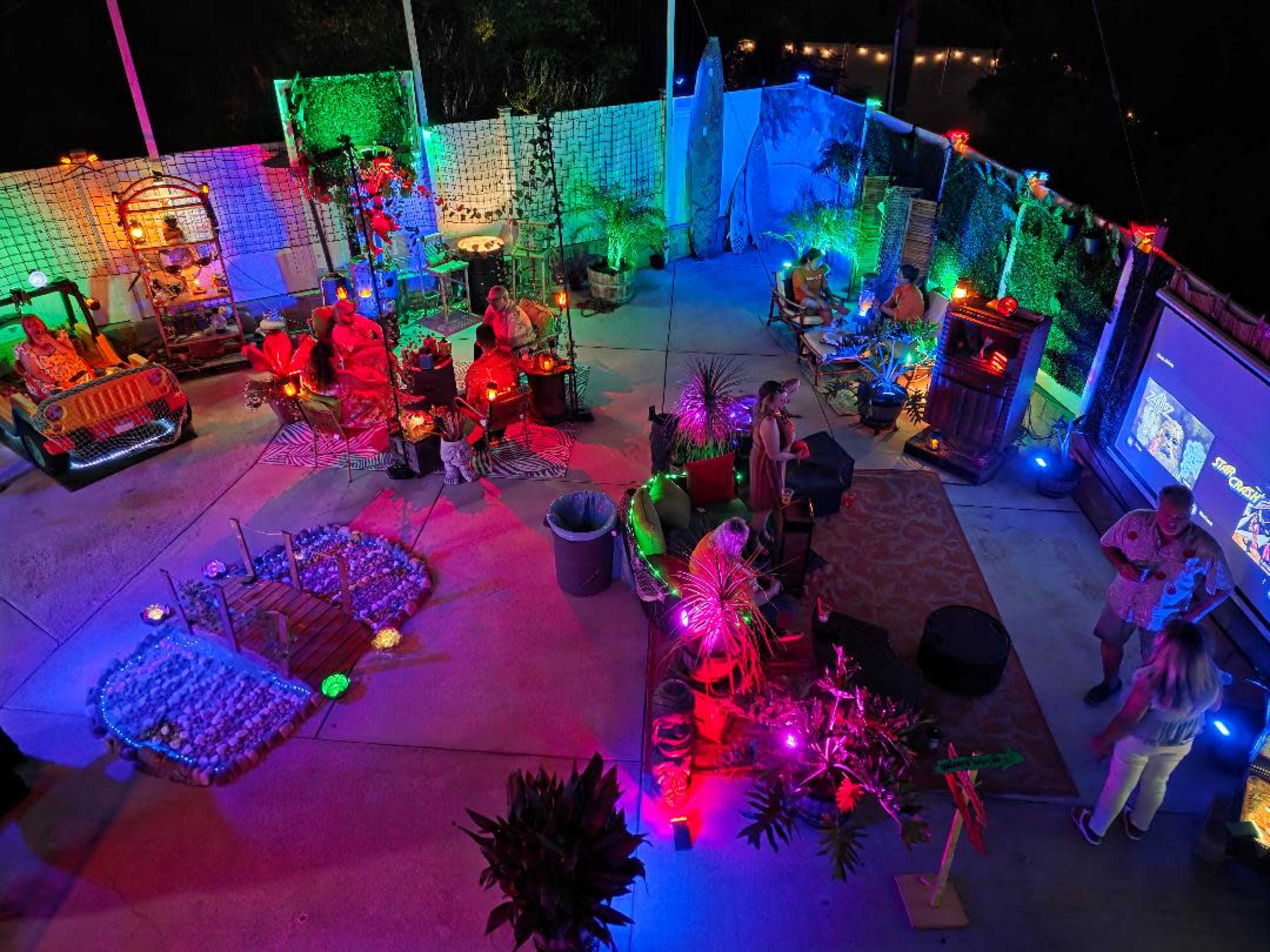 The scene shows an outdoor party area illuminated by vibrant colored lights, with guests socializing among various decorations and themed setups.