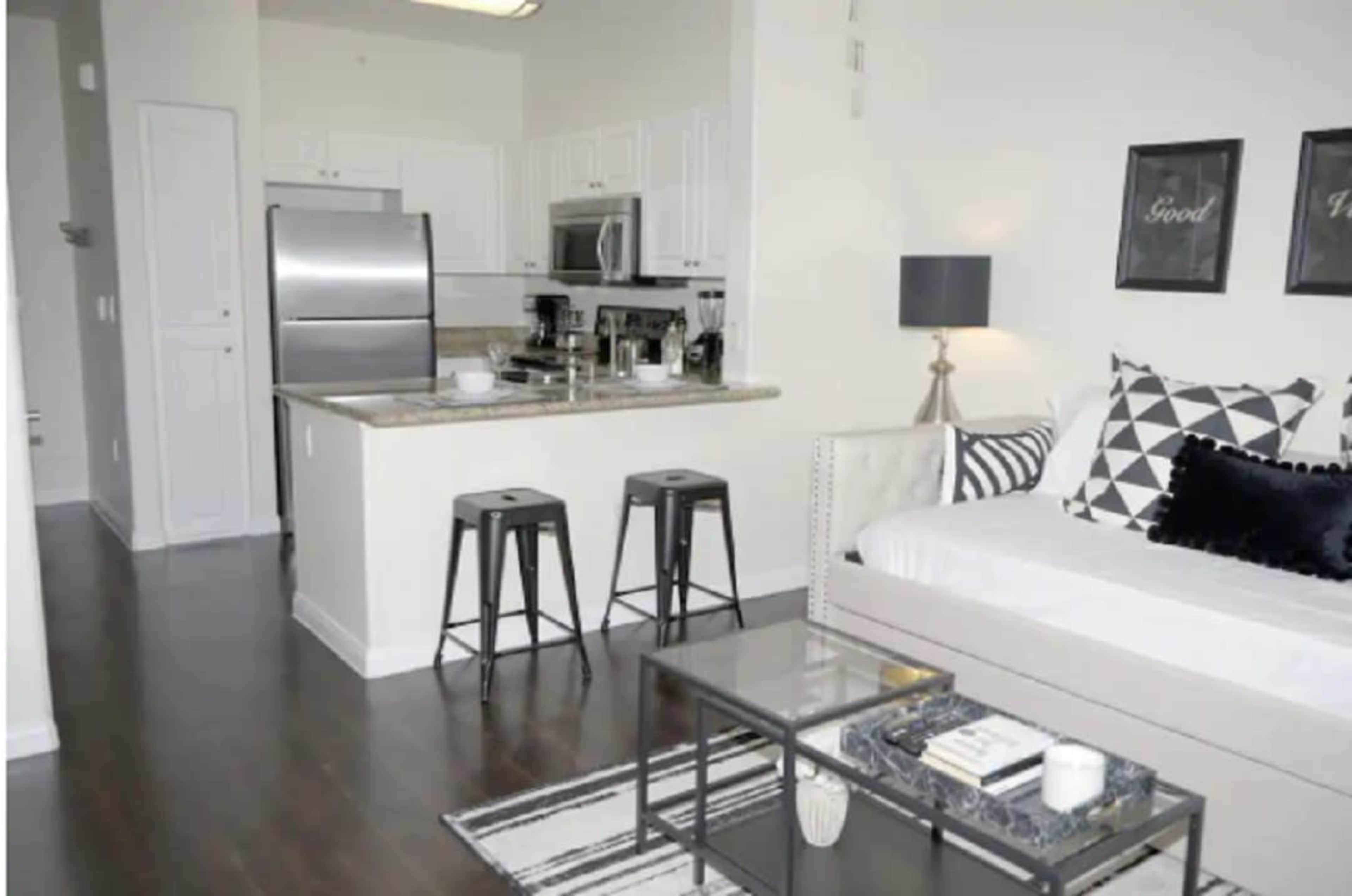 The image shows a modern, open-concept living space featuring a kitchen with stainless steel appliances, a sofa with decorative pillows, a coffee table, and bar seating.