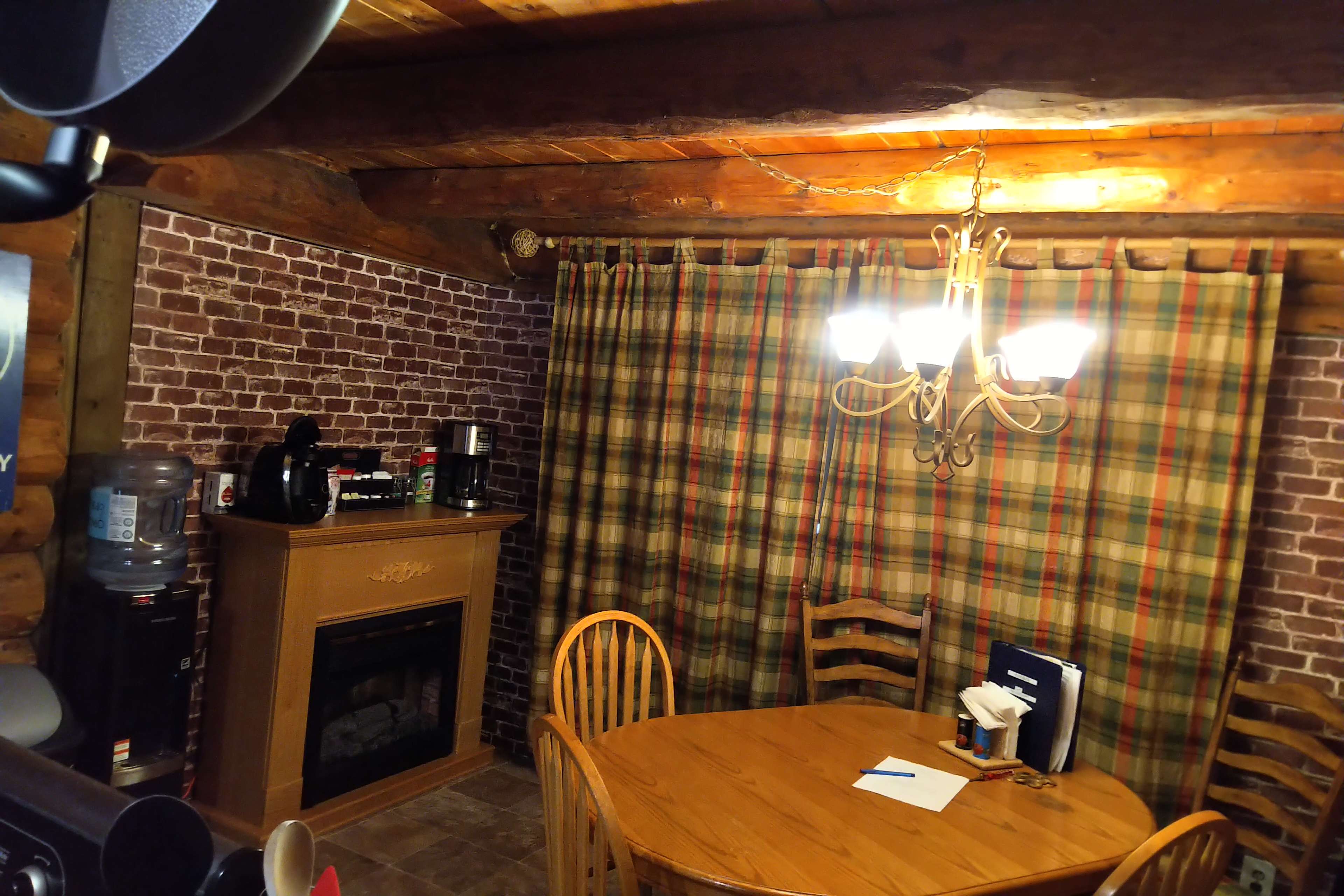 A cozy dining area with a wooden table, chairs, a plaid curtain, and a fireplace against a brick-patterned wall.