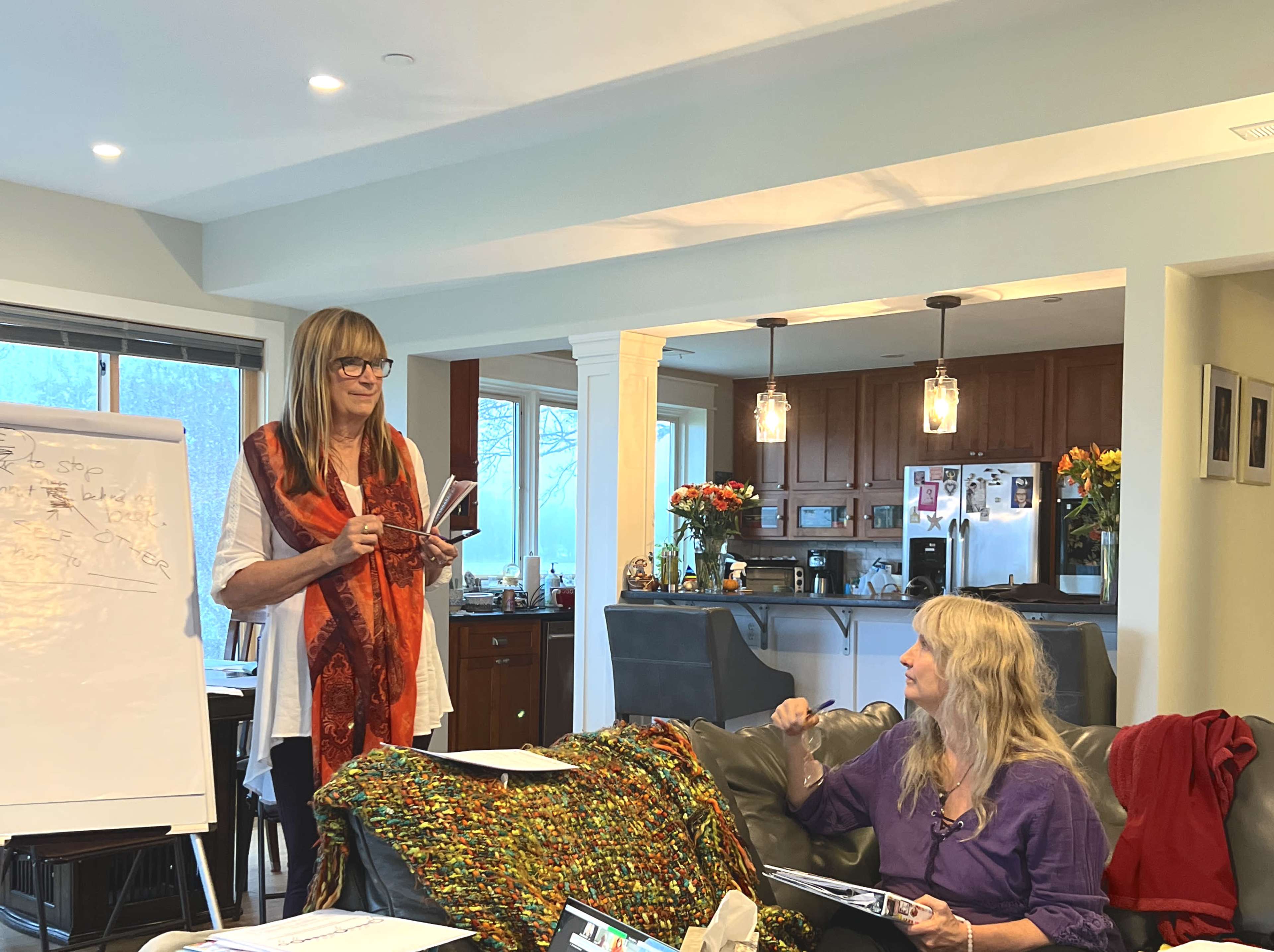 Two women participate in a meeting in a well-lit living room, with one presenting at a whiteboard and the other taking notes on a couch.