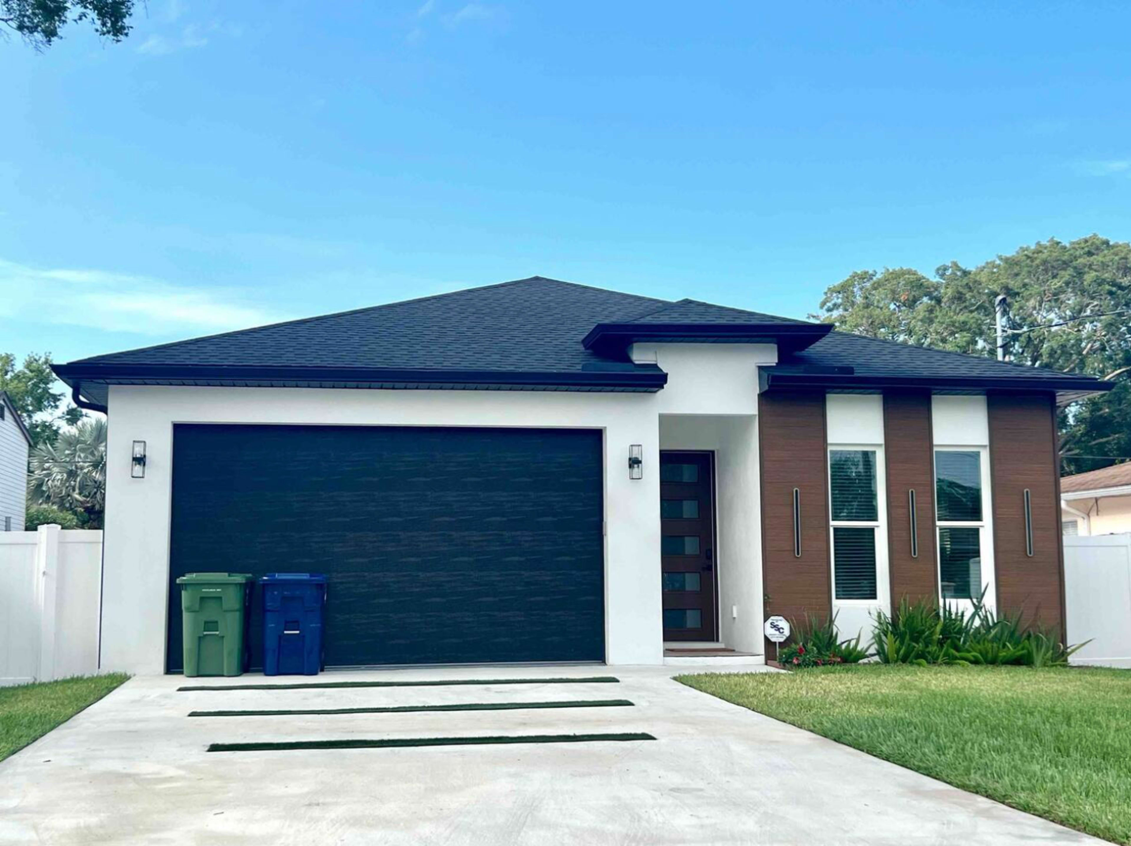A modern single-story house with a black garage door, two front windows, and a driveway lined with grass.