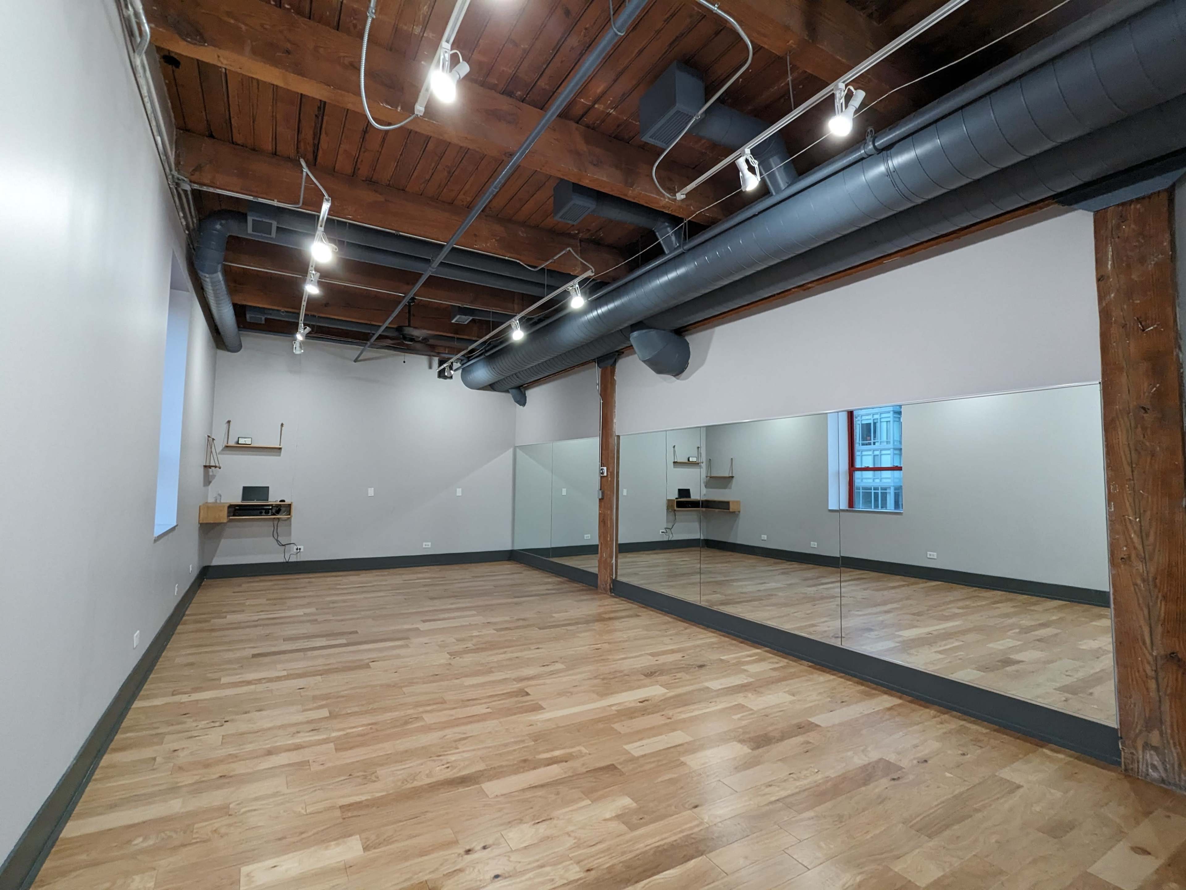 An empty studio space with wooden flooring, exposed wooden beams, and a large mirror along one wall.