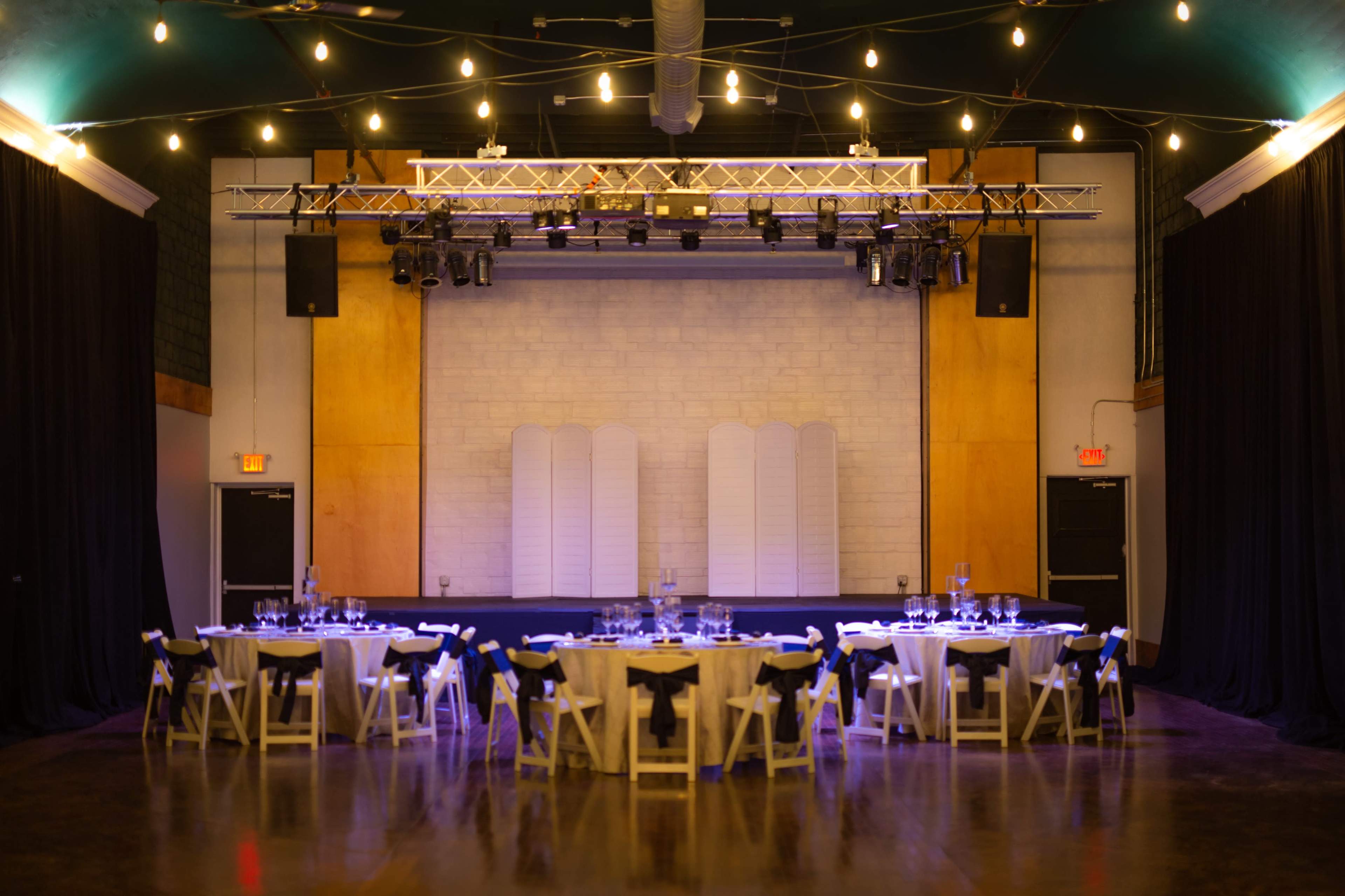 A banquet hall is set up with circular tables arranged for dining, featuring white tablecloths and chairs, under decorative string lights and a stage in the background.