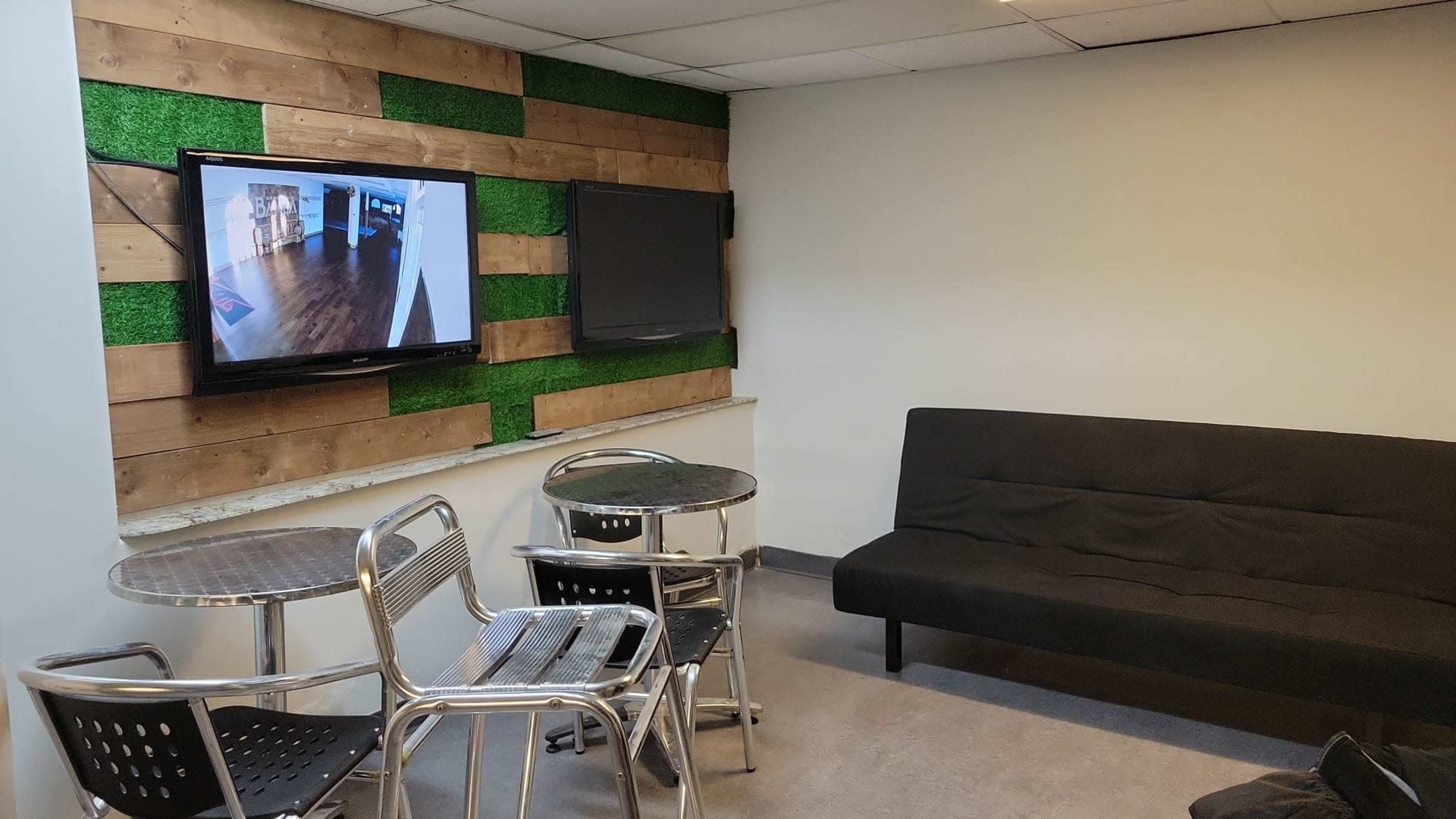 A room features a black sofa and two round metal tables with chairs, along with two wall-mounted televisions and a wooden accent wall.