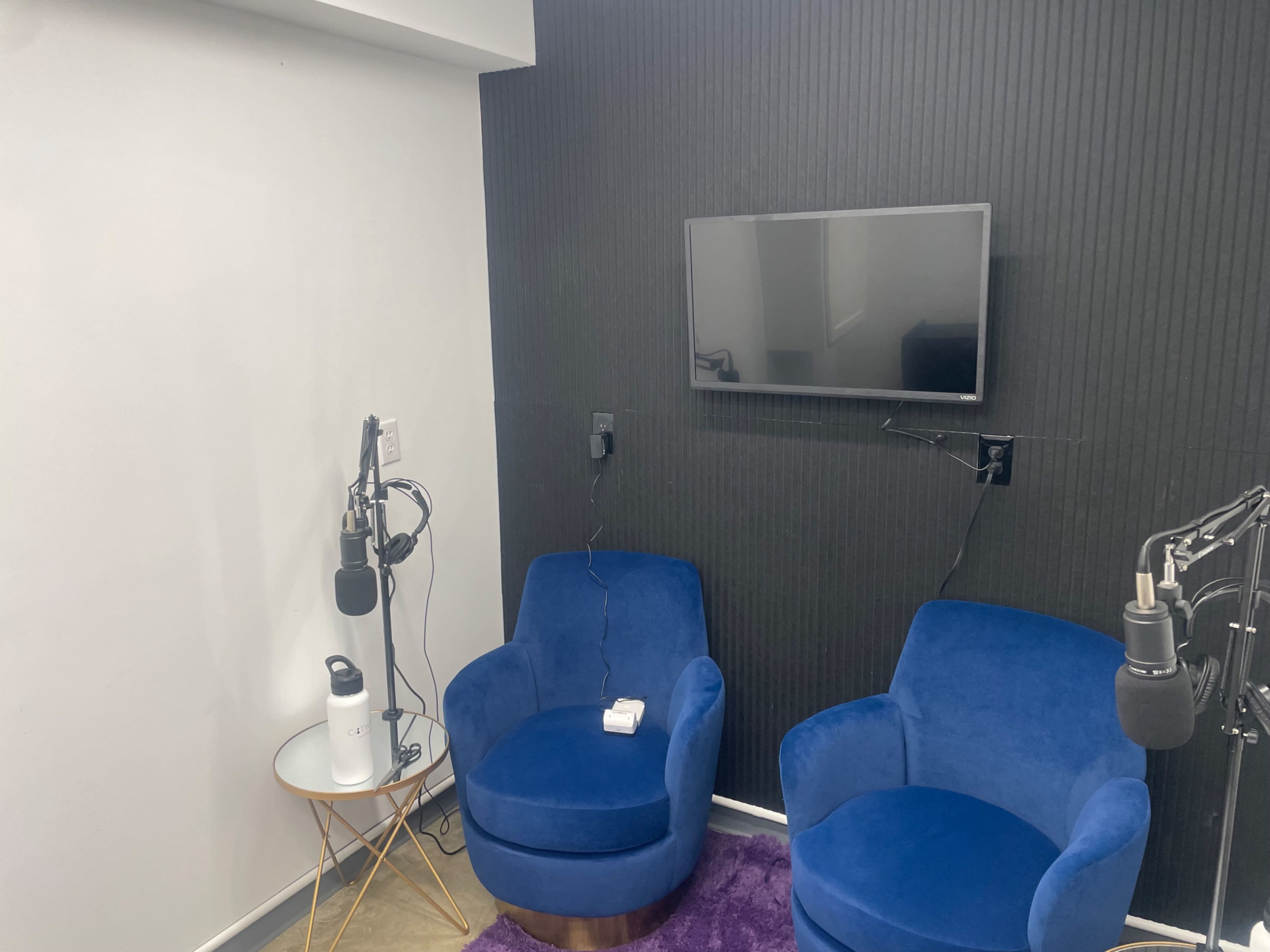 The image shows a small recording studio with two blue chairs, a coffee table, and a wall-mounted television, equipped with microphones.