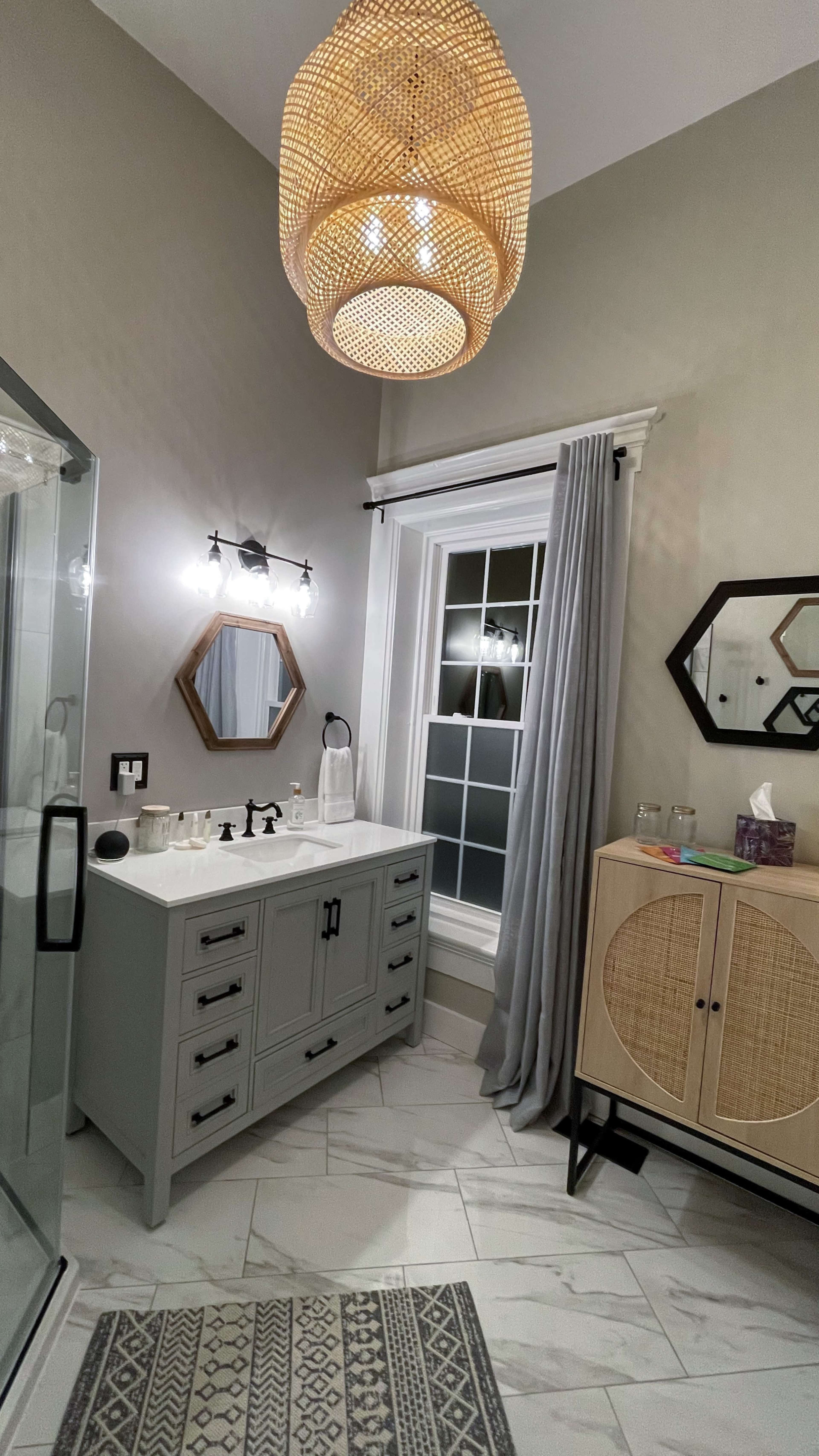 A modern bathroom features a gray vanity, a glass shower, and a woven light fixture above, with large windows and geometric mirrors on the walls.