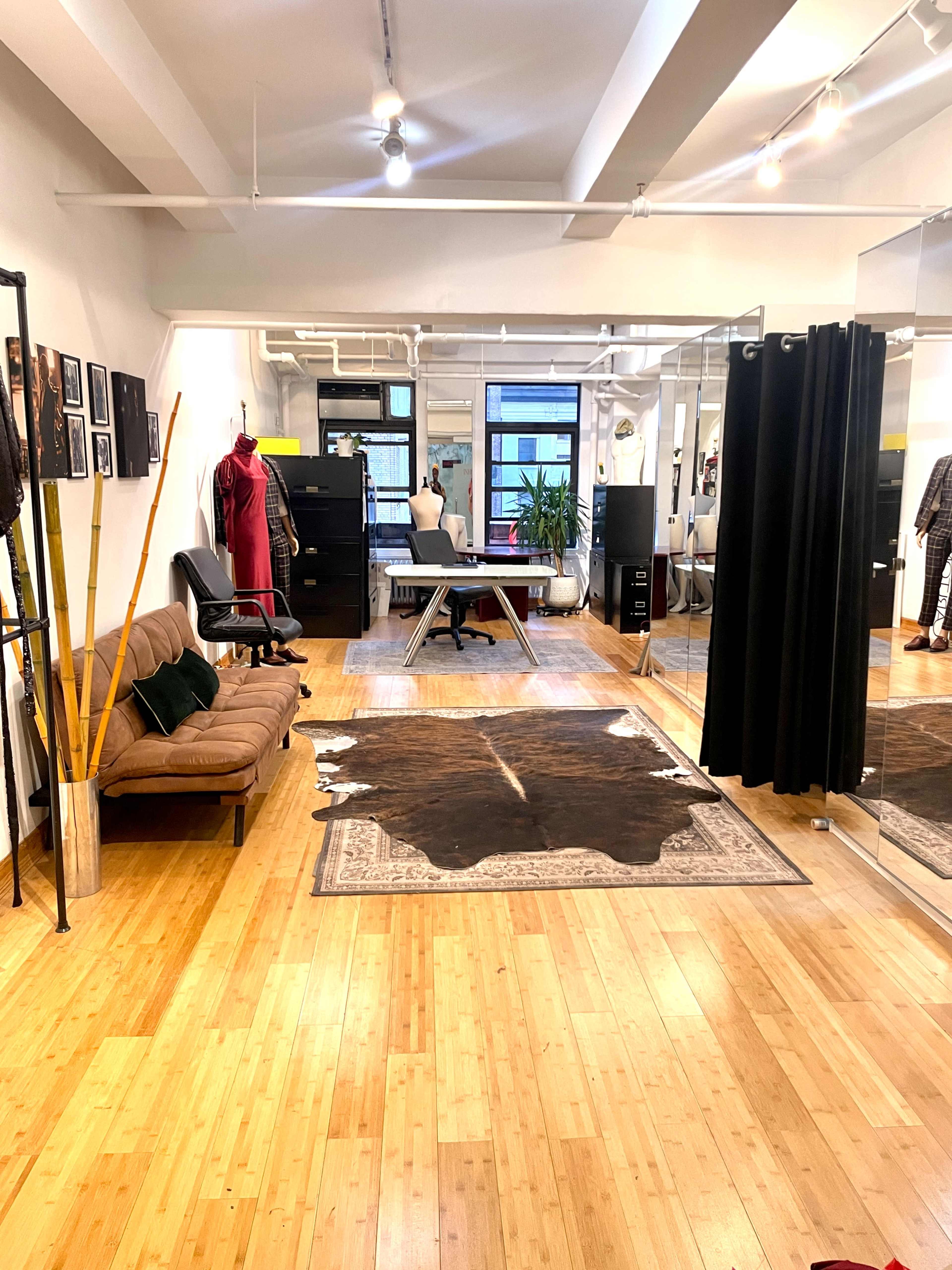 The image shows a spacious studio with wooden floors, a brown sofa, a large mirror, a black curtain, a tailored work table, and a decorative cowhide rug.