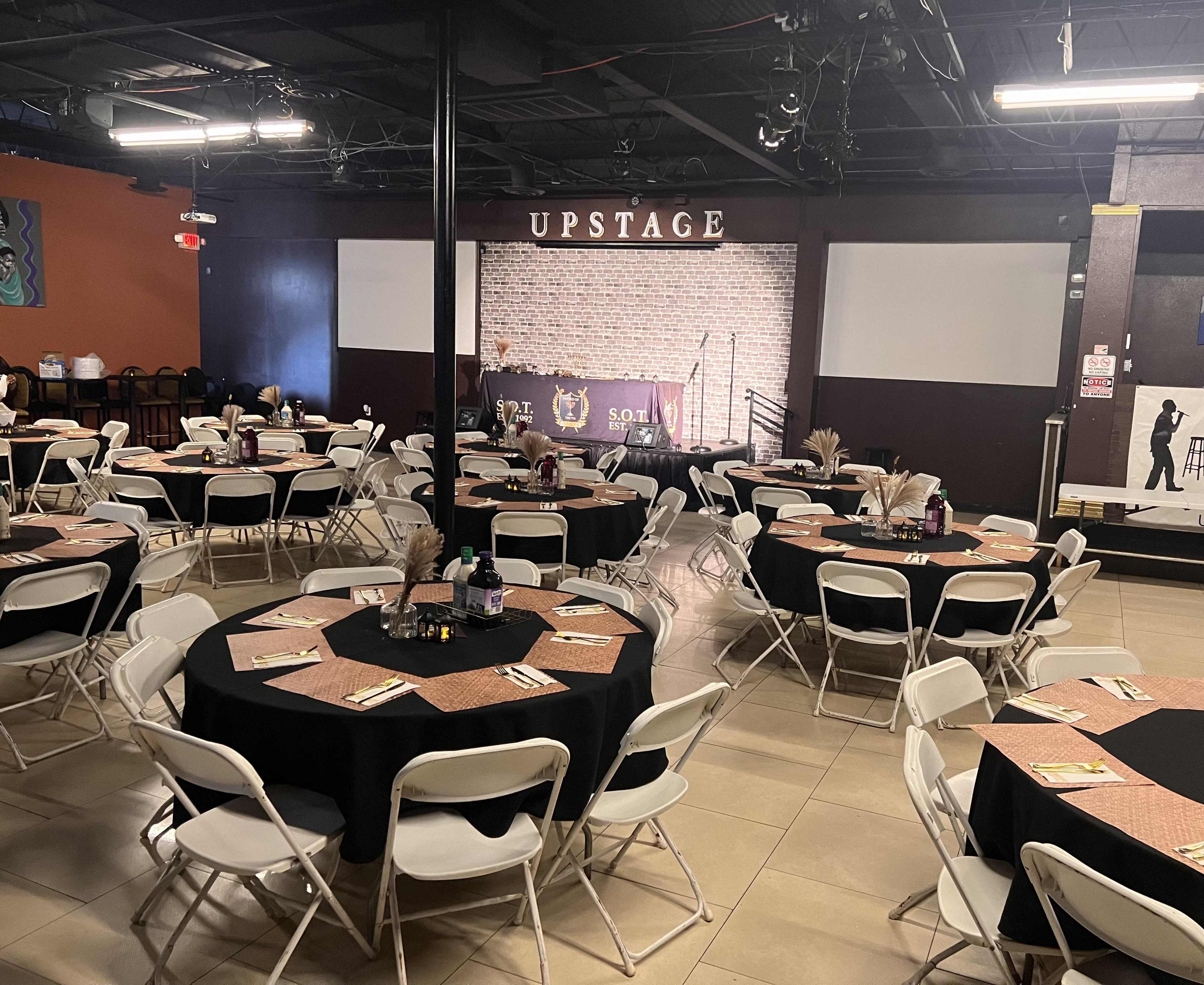 The image shows a spacious event venue with round tables set for dining, featuring black tablecloths and centerpieces, and a stage area with a backdrop labeled "UPSTAGE."