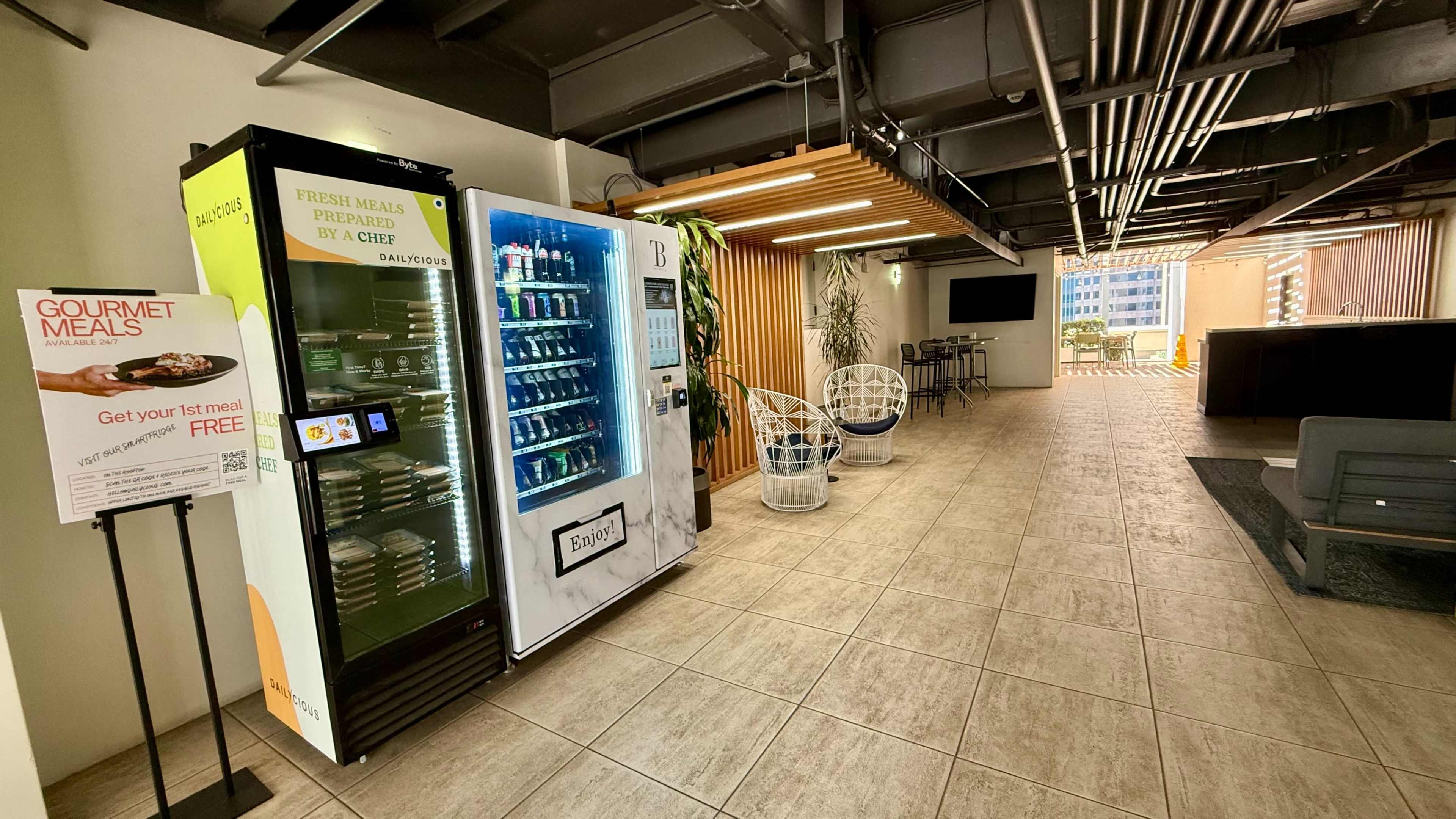 The image shows a modern hallway featuring a vending machine with fresh meals, a seating area, and an open space with natural light coming from large windows.