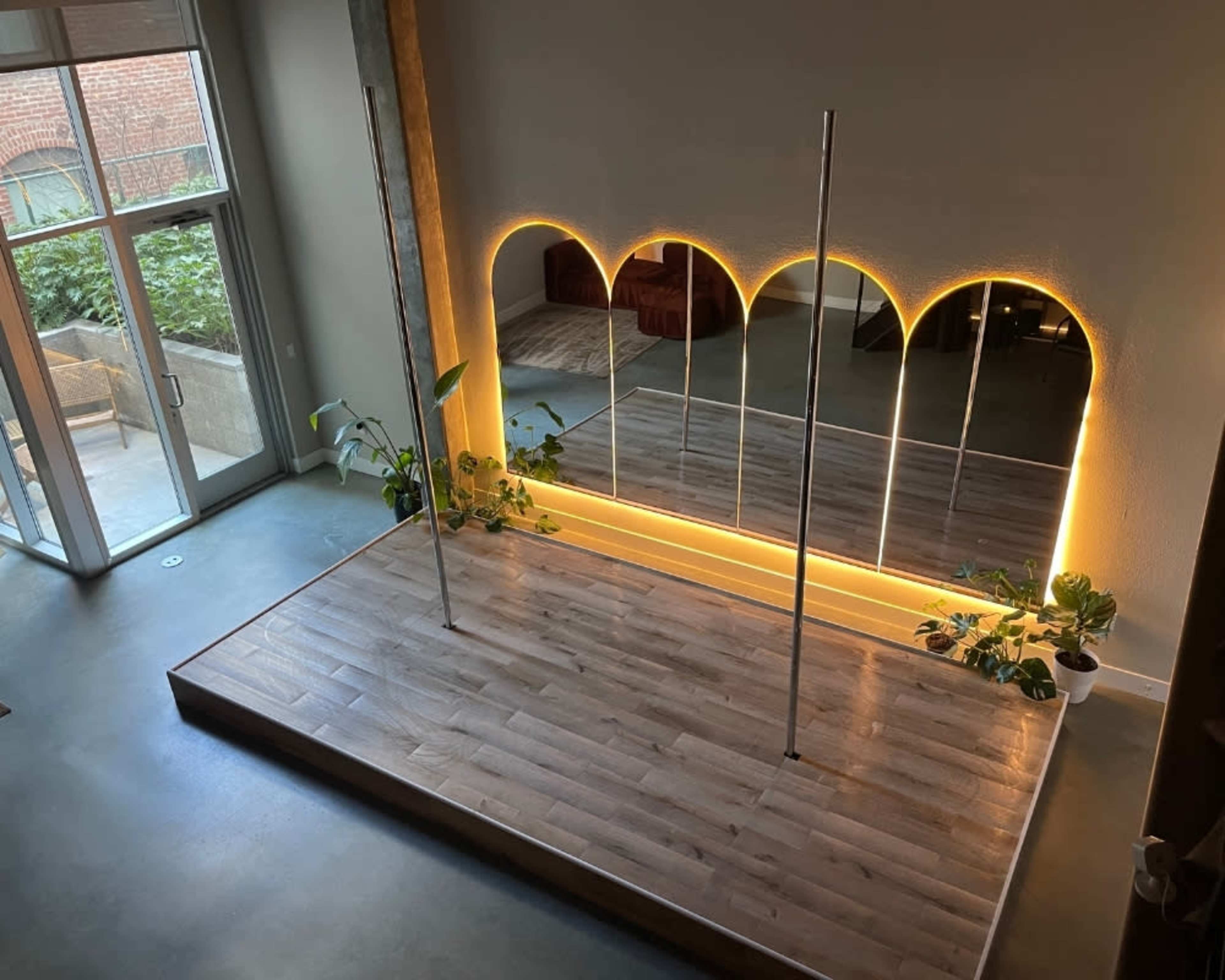 The image shows a spacious dance studio with a wooden floor, two dance poles, and a wall of mirrors illuminated by soft lighting.