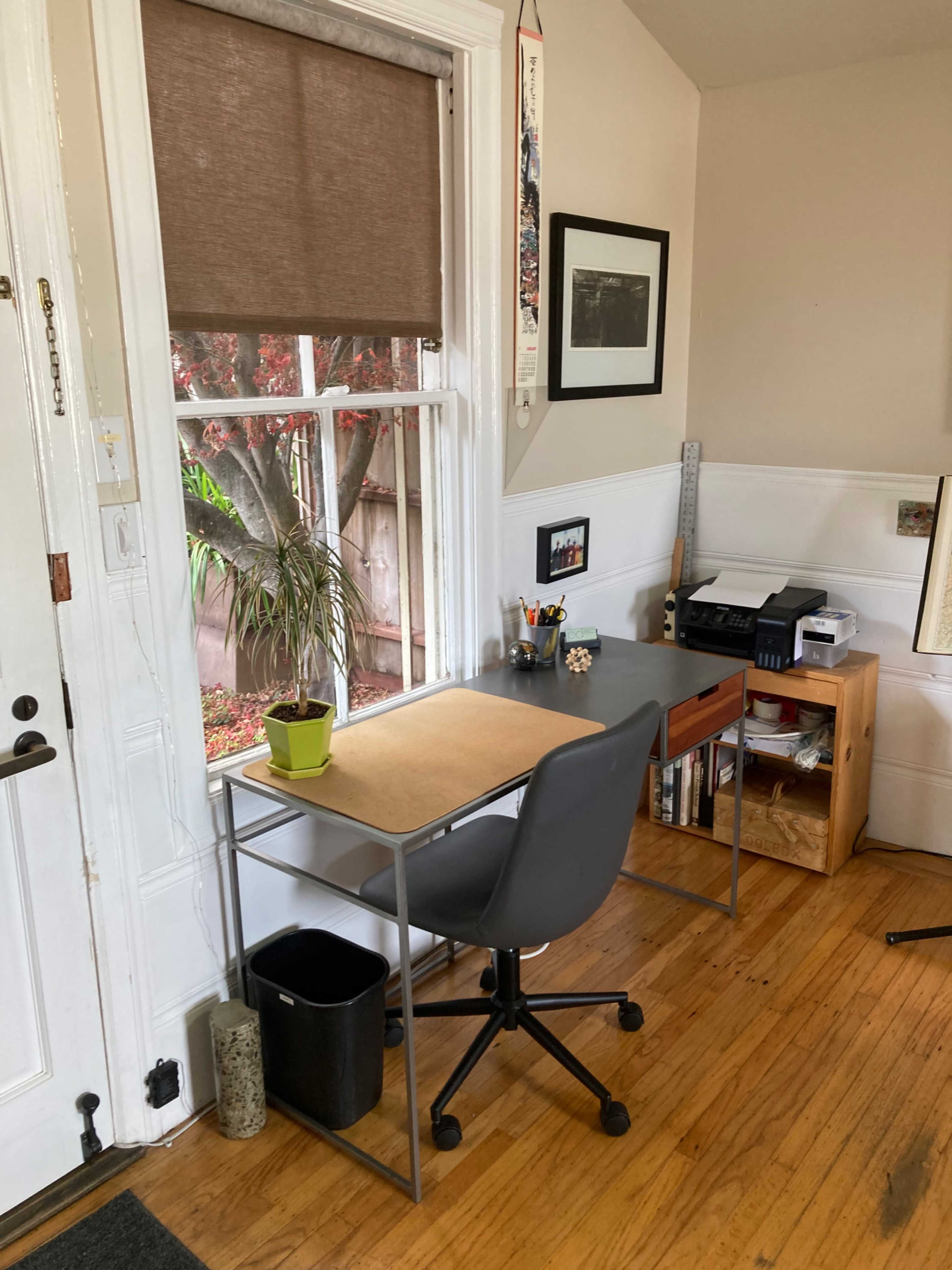A small home office features a desk with a chair, a window with a view of greenery, and storage furniture against the wall.