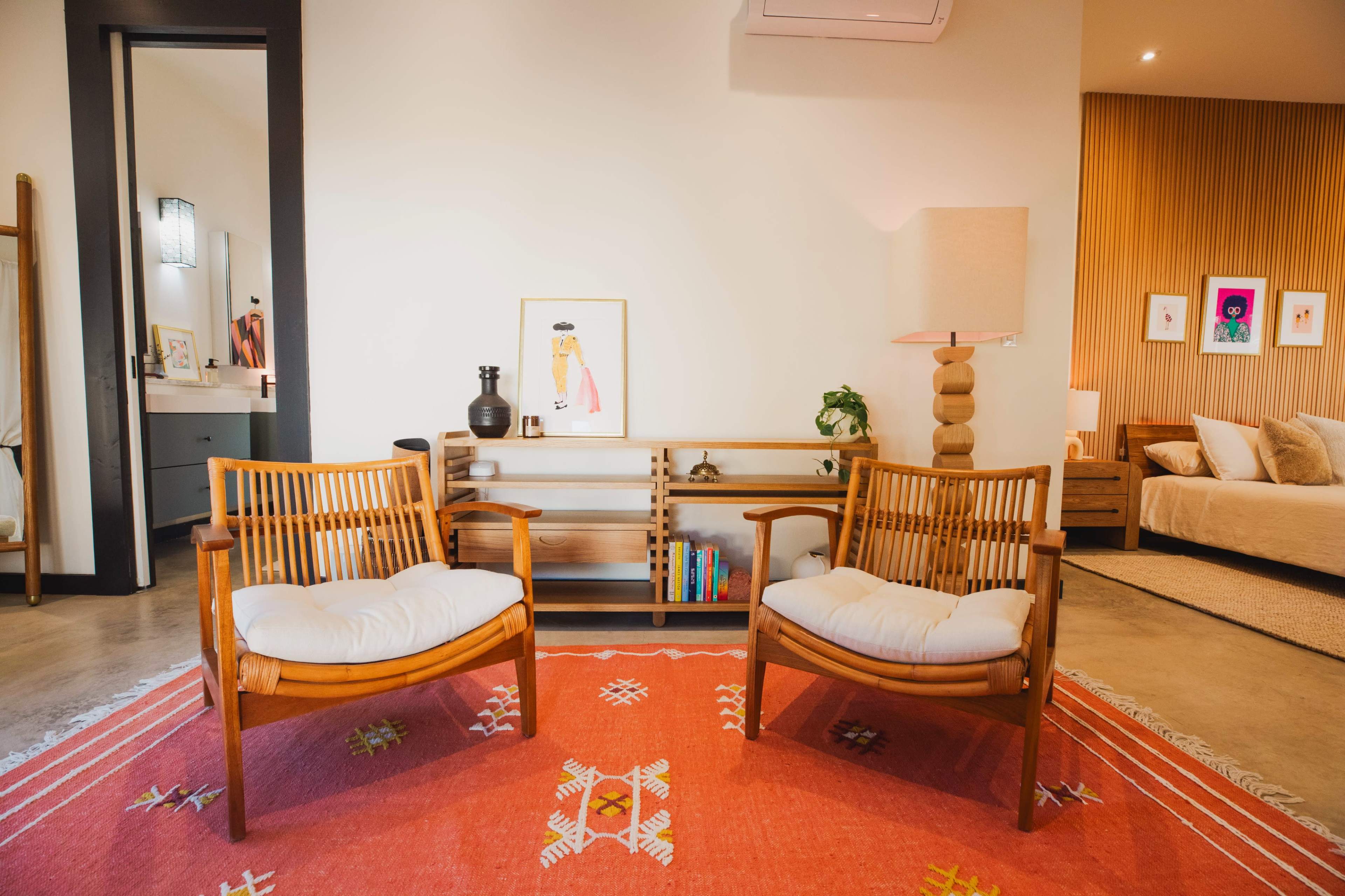 A modern living space featuring two wooden lounge chairs with cushions, a colorful area rug, a small side table with a lamp, and a decorative wall in the background.