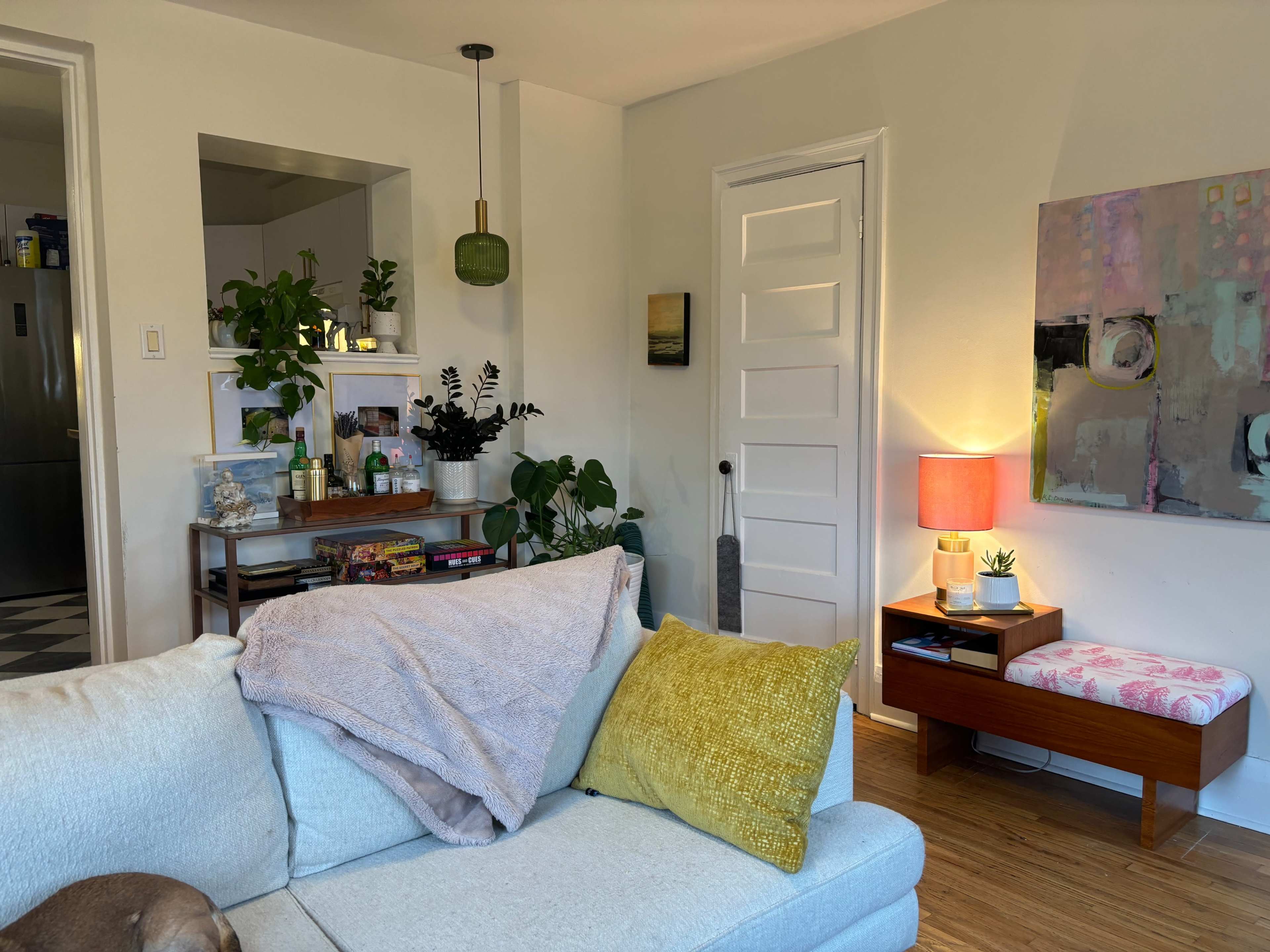 A cozy living room with a light blue sofa, a yellow throw pillow, a small wooden table with a pink lamp, and various plants on display.
