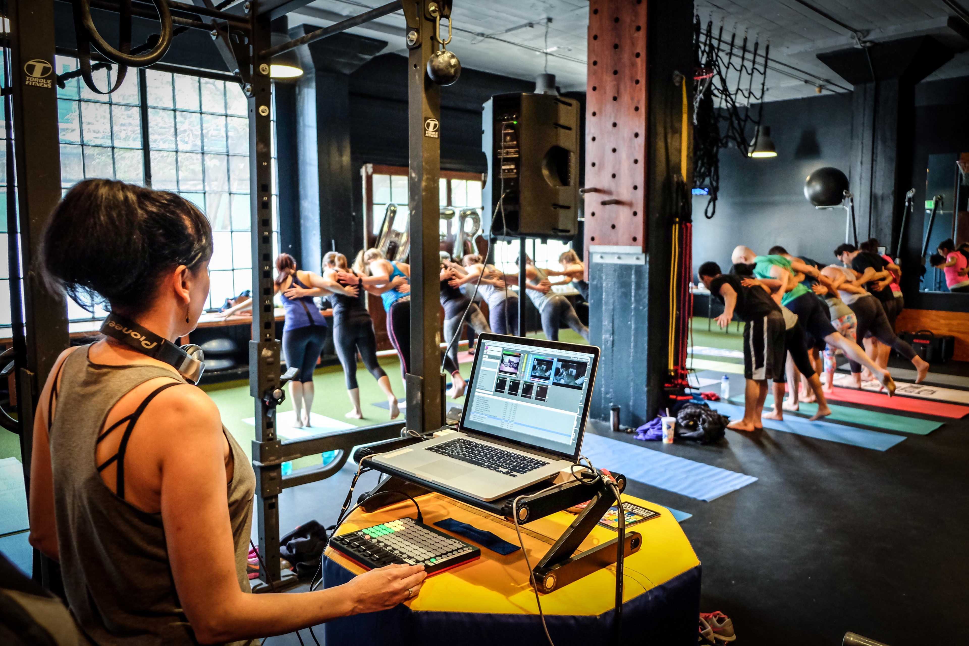 A fitness instructor monitors a group workout session while using a computer and sound equipment at the front of the gym.