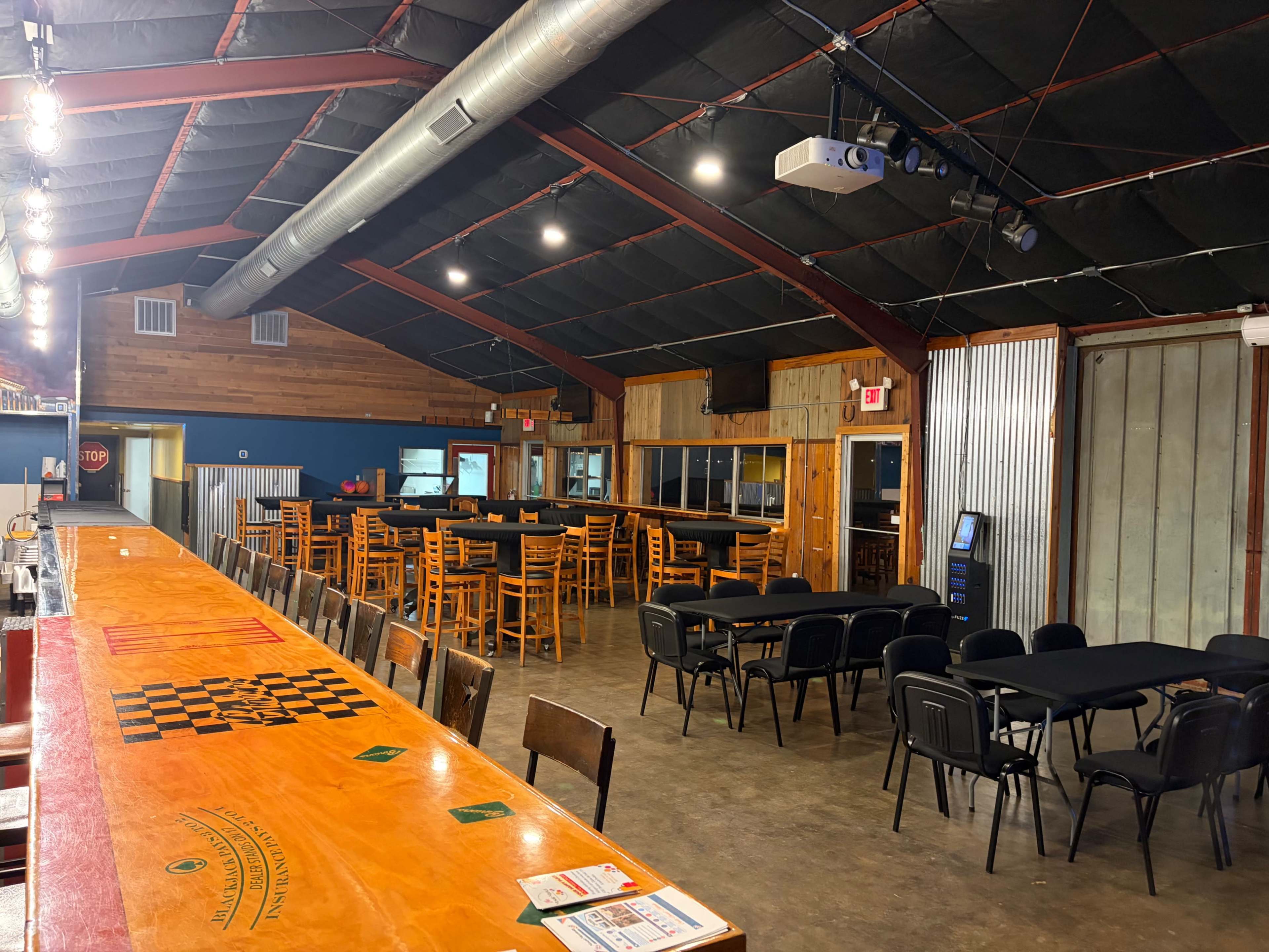 The interior of a rustic venue features a long wooden bar and multiple dining tables arranged in a spacious room with exposed metal and wooden accents.