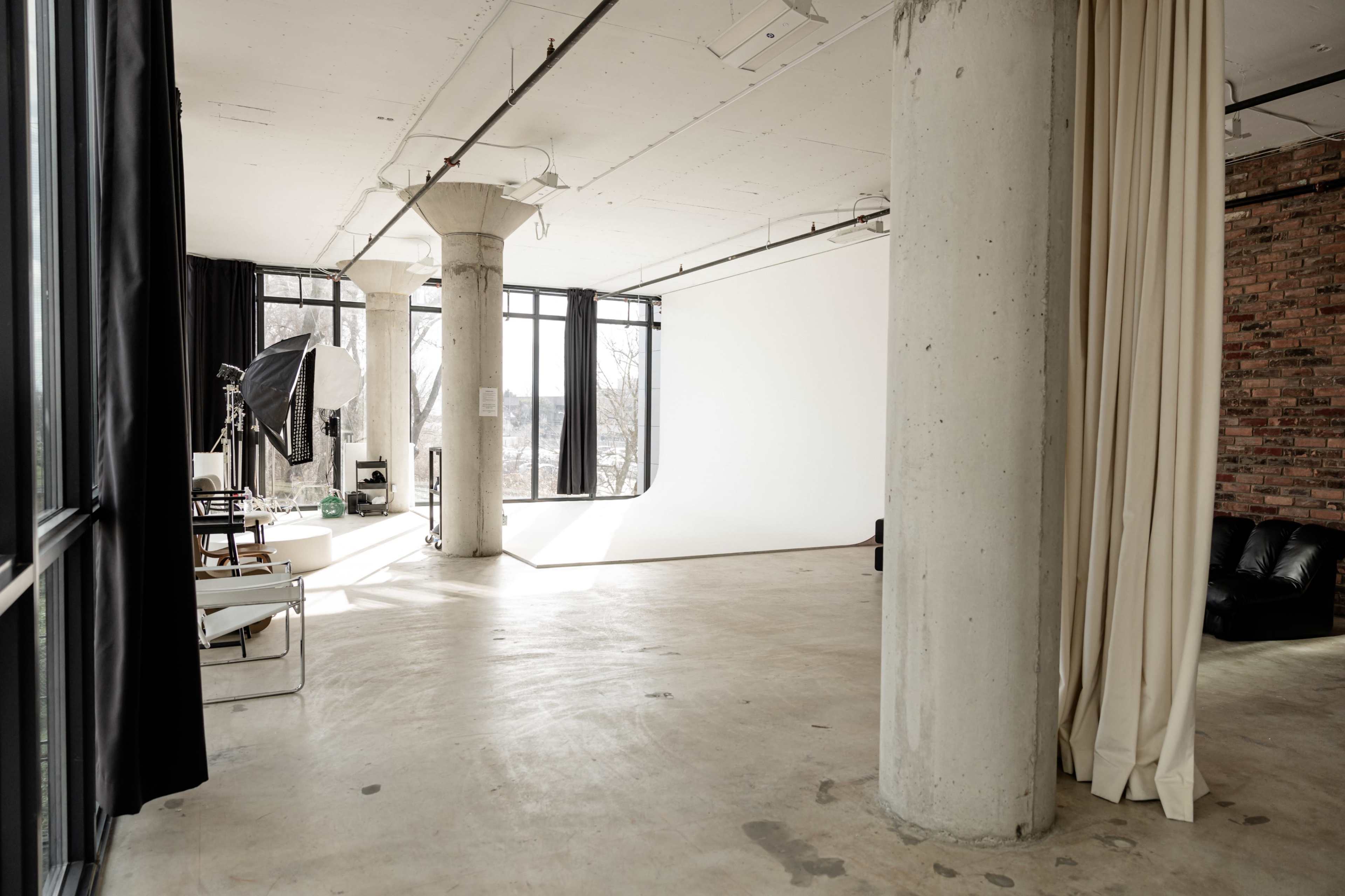 The image shows a spacious photography studio with large windows, concrete pillars, and a seamless white backdrop.