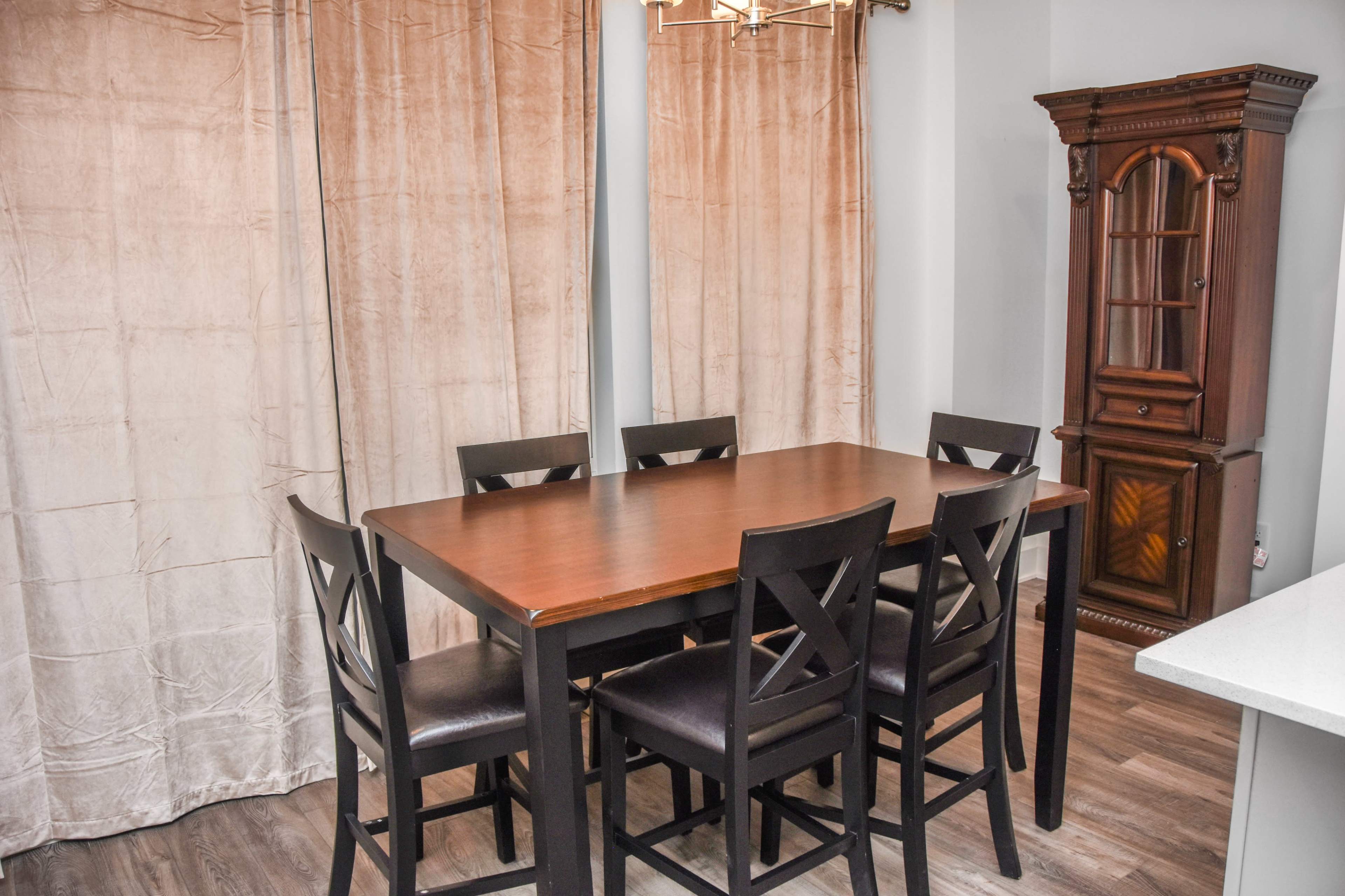 A wooden dining table surrounded by six black chairs is positioned in a well-lit room with beige curtains and a wooden cabinet in the corner.