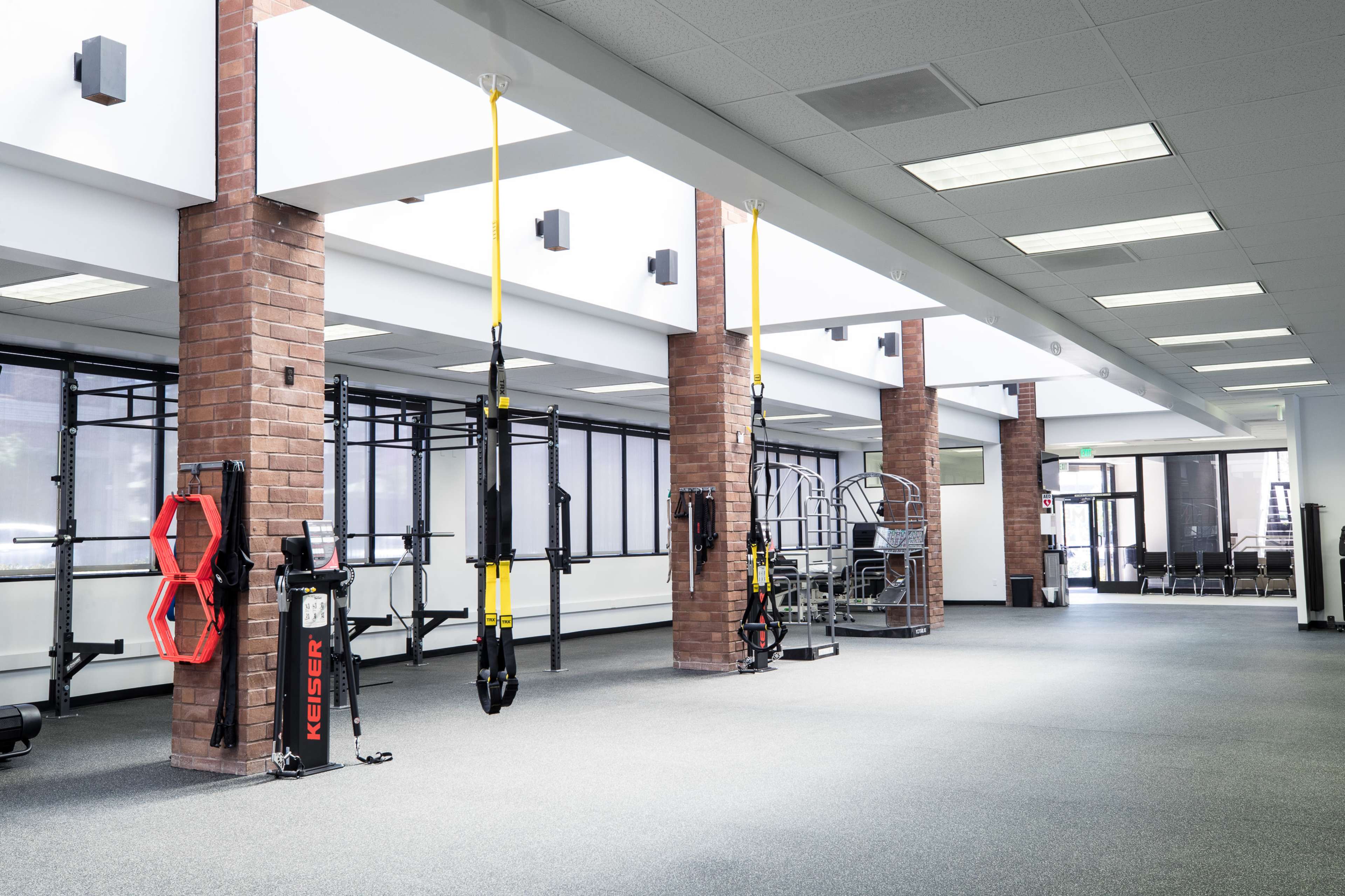 The image shows a modern gym interior featuring various exercise equipment, including suspension trainers and a multi-station weight machine, in an open layout with brick columns.