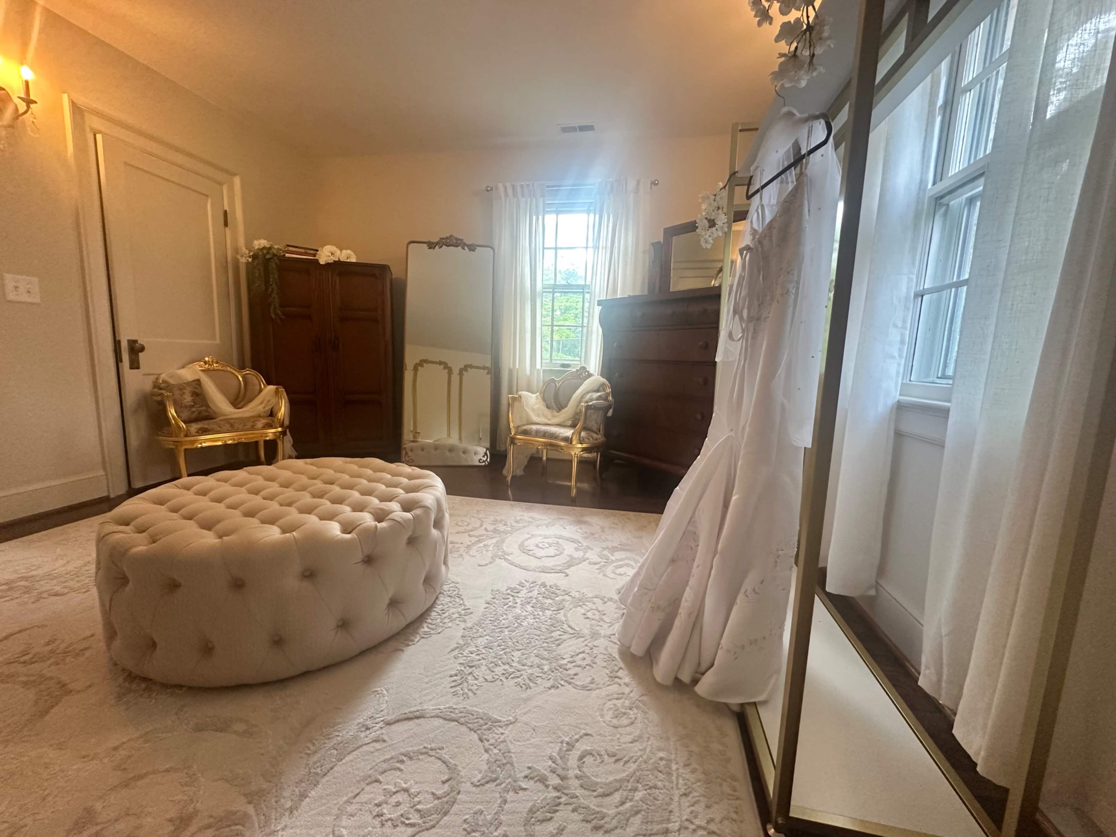 The image shows a softly lit dressing room featuring a large tufted ottoman, a wedding dress hanging on a mirror, and elegant furniture, including a mirrored wardrobe and two gold chairs.