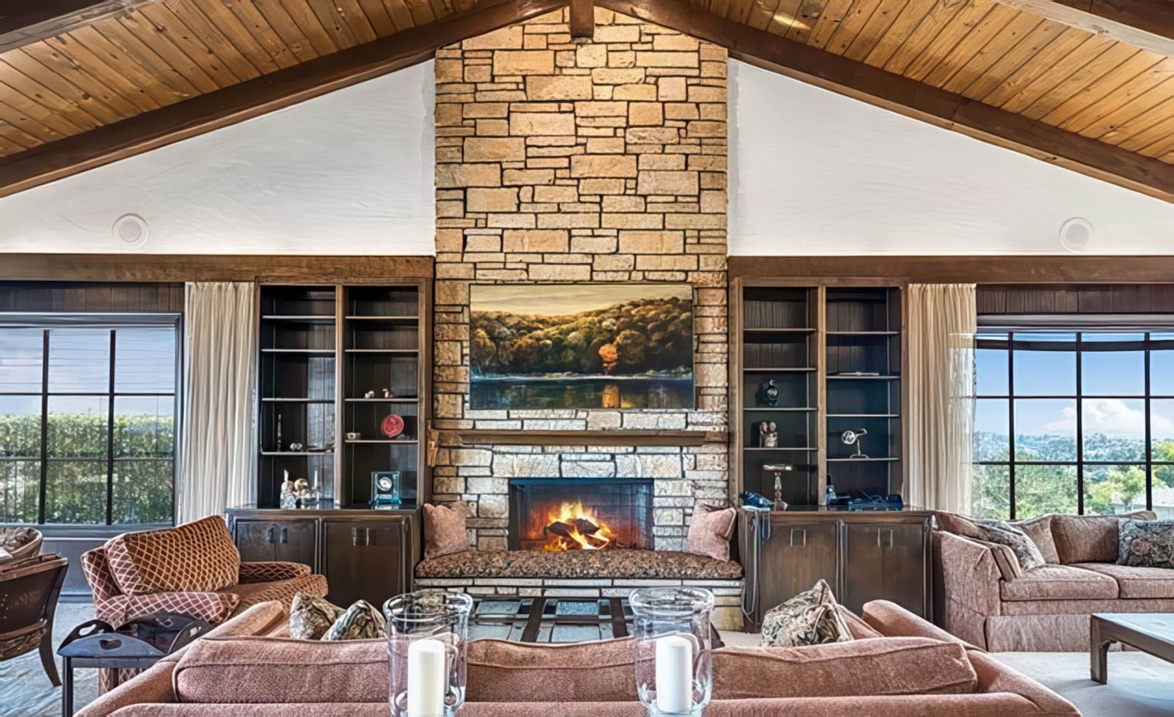 The image features a spacious living room with a stone fireplace, large windows, and wooden beams on the ceiling, surrounded by comfortable seating and built-in shelves.