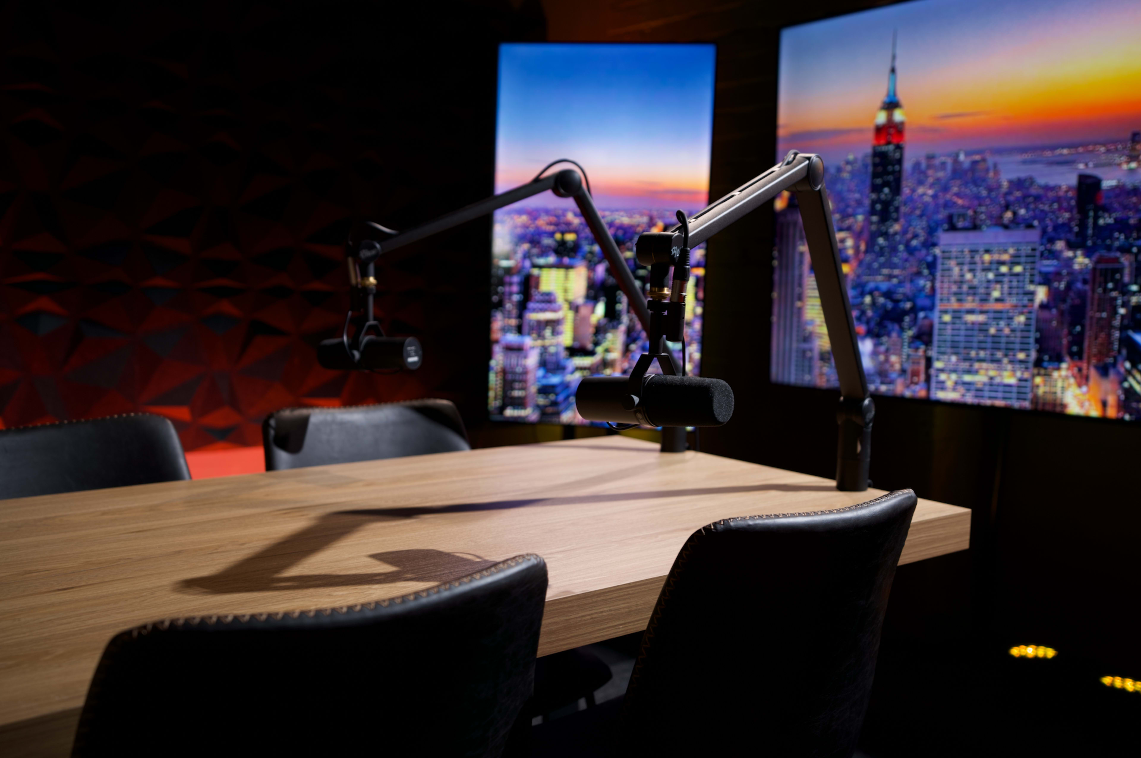 A wooden table and four chairs are set up in a recording studio with two large screens displaying a city skyline.