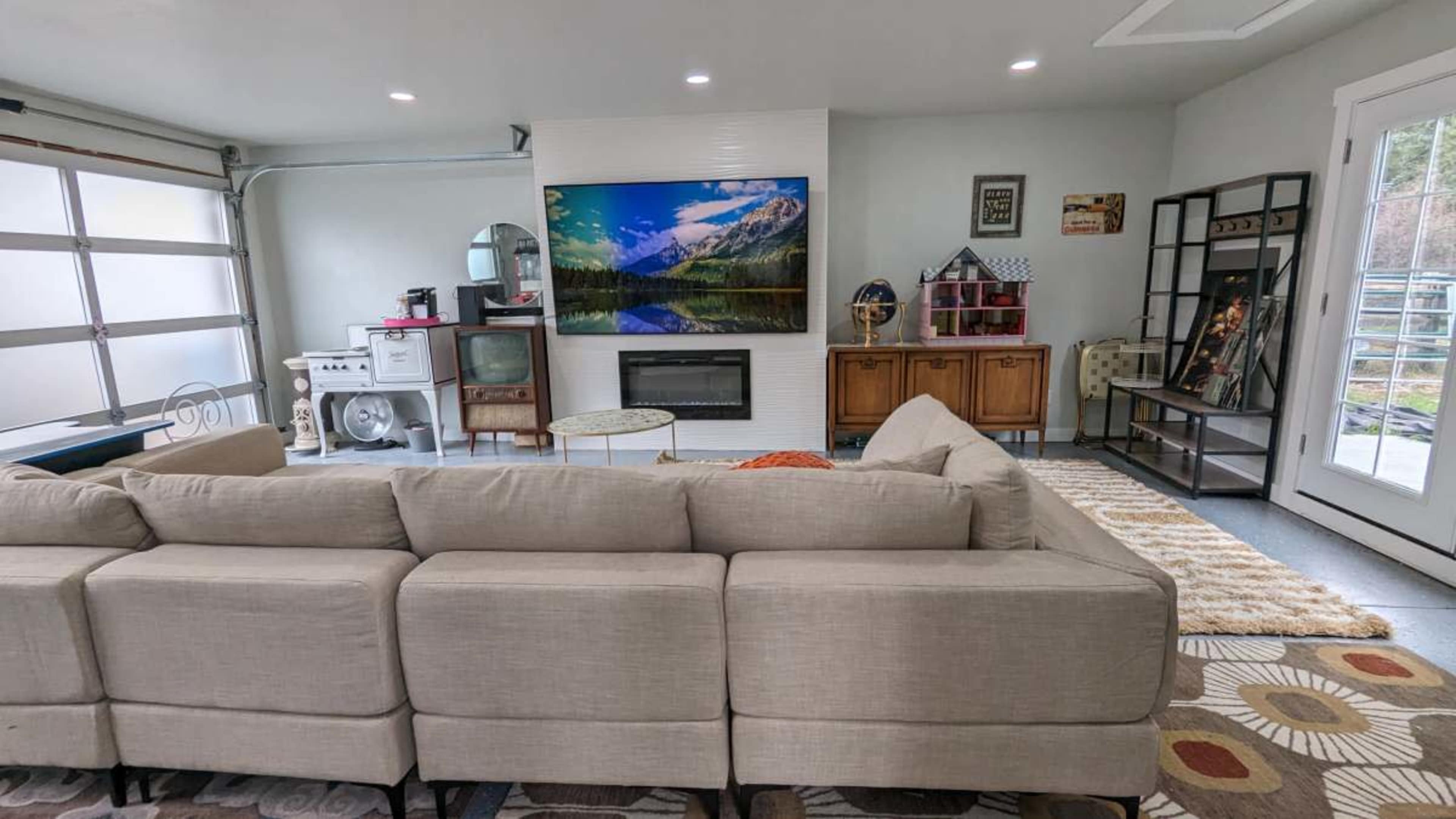 The image shows a modern living room featuring a beige sectional sofa facing a wall-mounted television above an electric fireplace, with a mix of furniture and decor throughout the space.