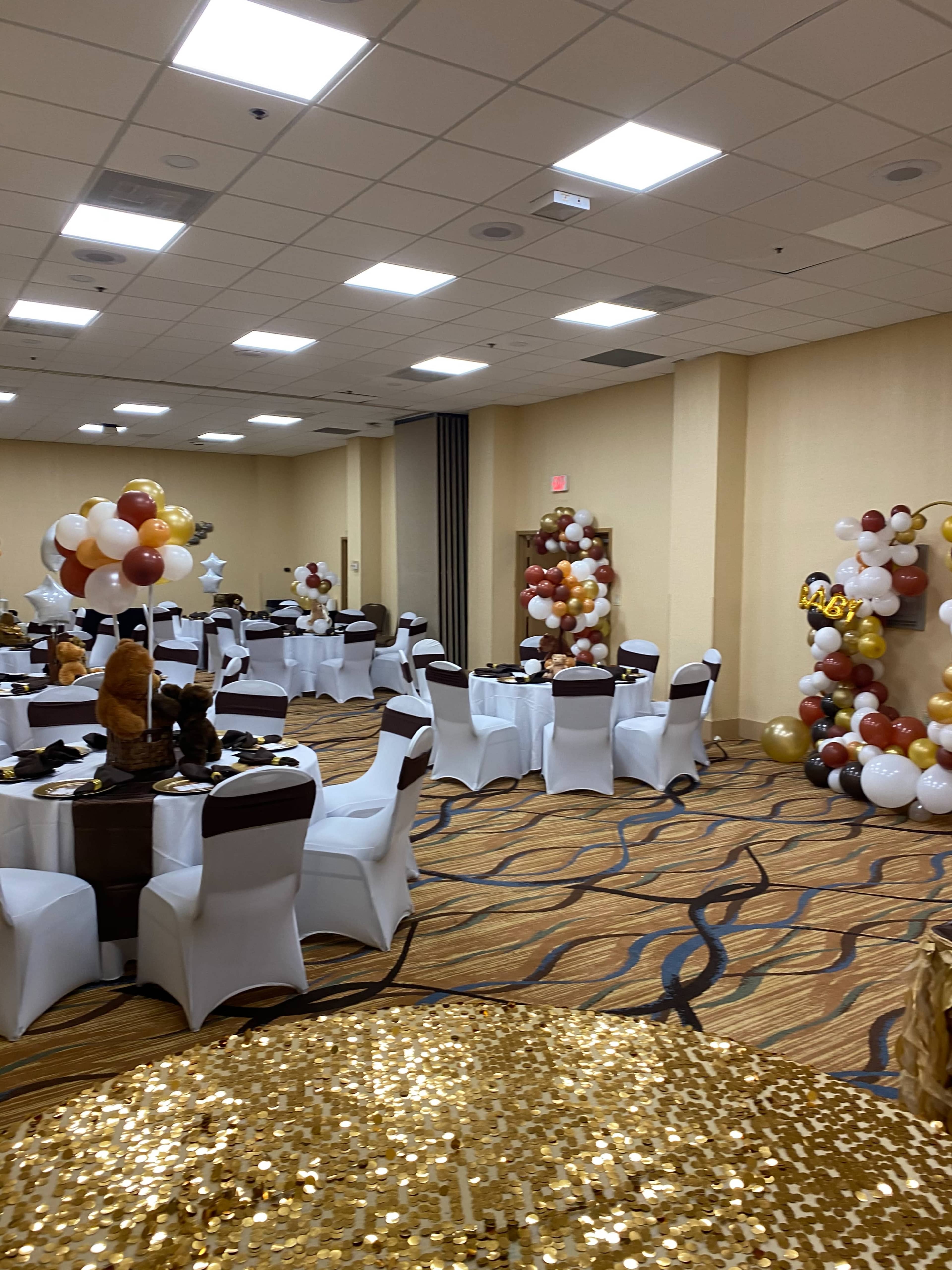 A banquet hall set for an event, featuring round tables covered with white tablecloths and decorated with balloons and gold accents.