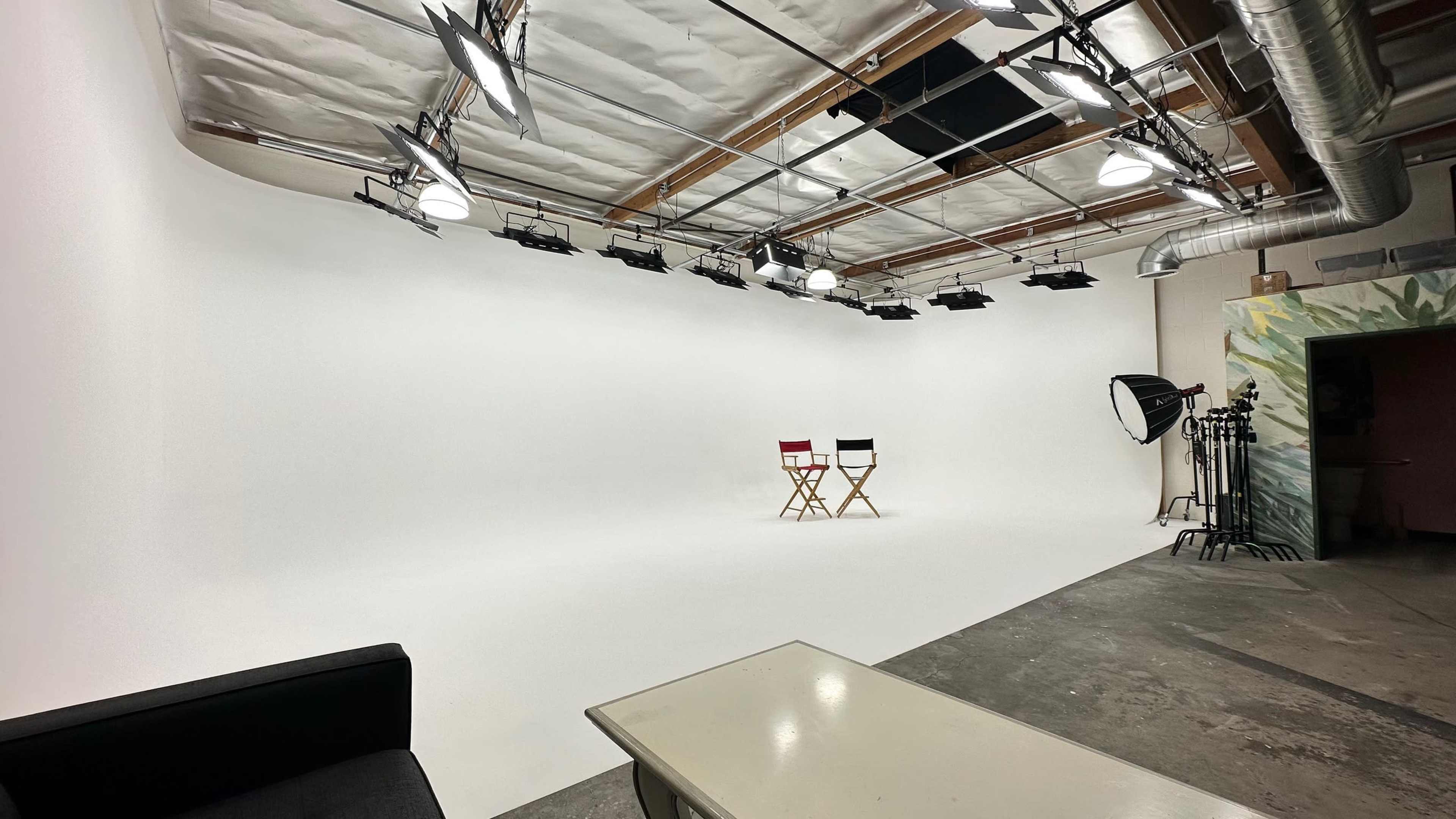 A photography studio with a white backdrop, two director's chairs, and various lighting equipment set up nearby.