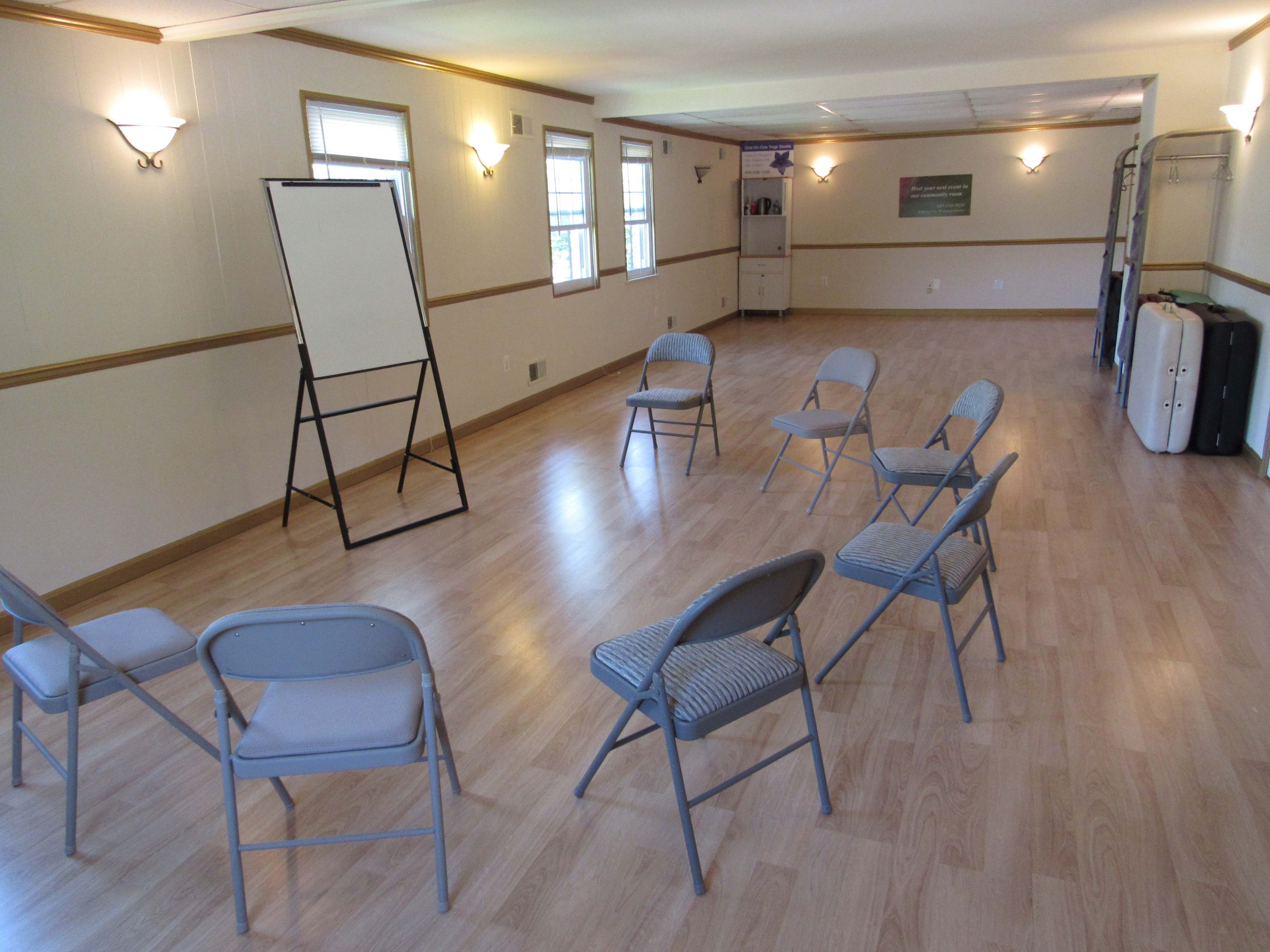 A room with a light wooden floor, featuring a set of six gray folding chairs arranged in a circle around a blank presentation board on an easel.