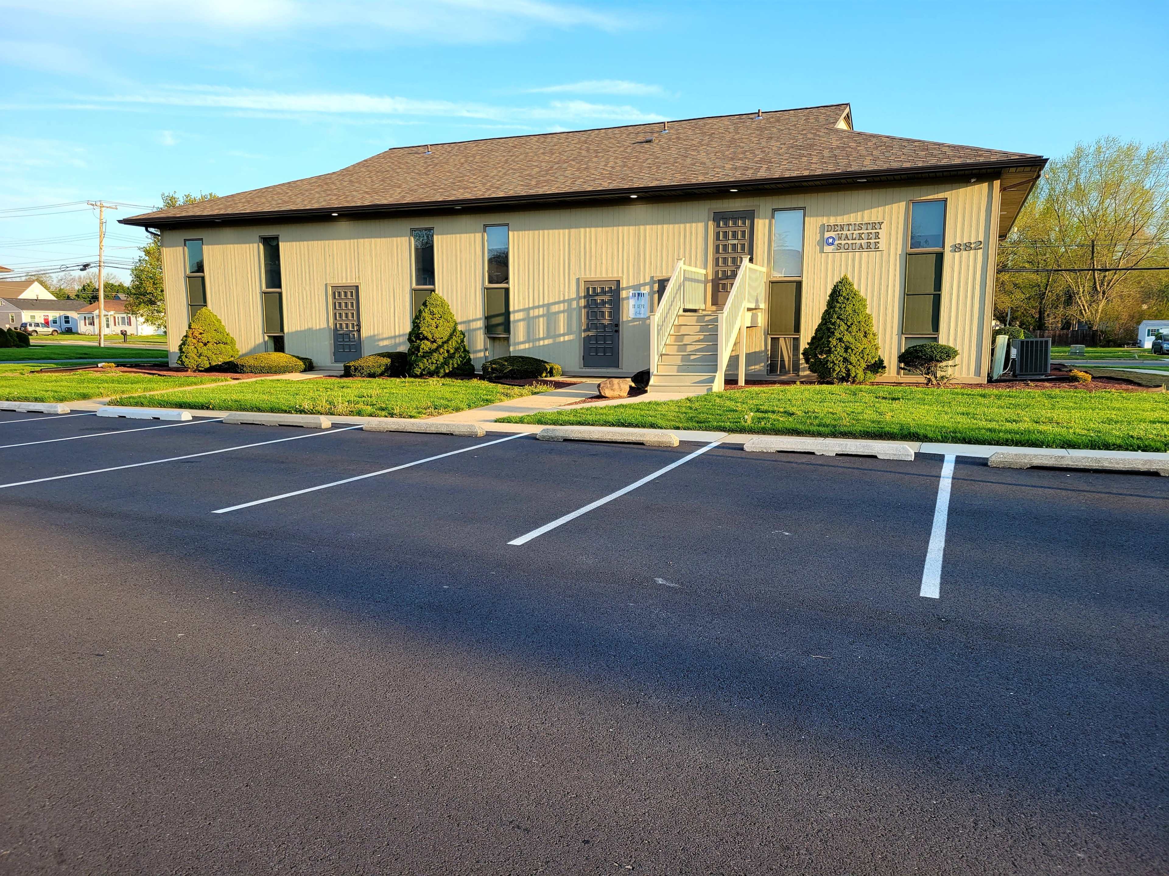 A two-story beige building with a sloped roof, surrounded by neatly trimmed shrubs and a parking lot with empty spaces in front.