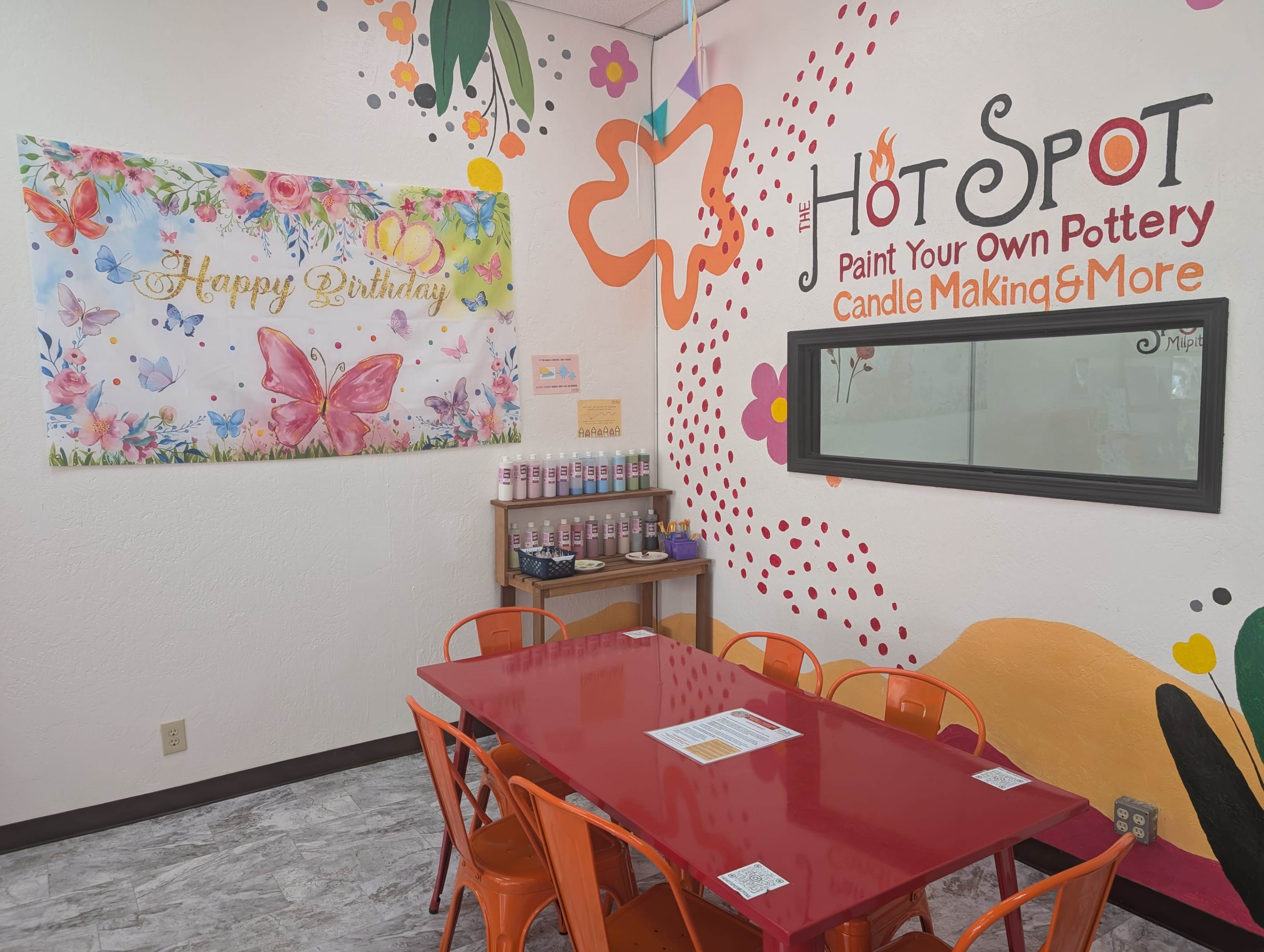 The image shows a brightly decorated art studio featuring a red table surrounded by orange chairs, with a colorful birthday banner and various pottery supplies on display.