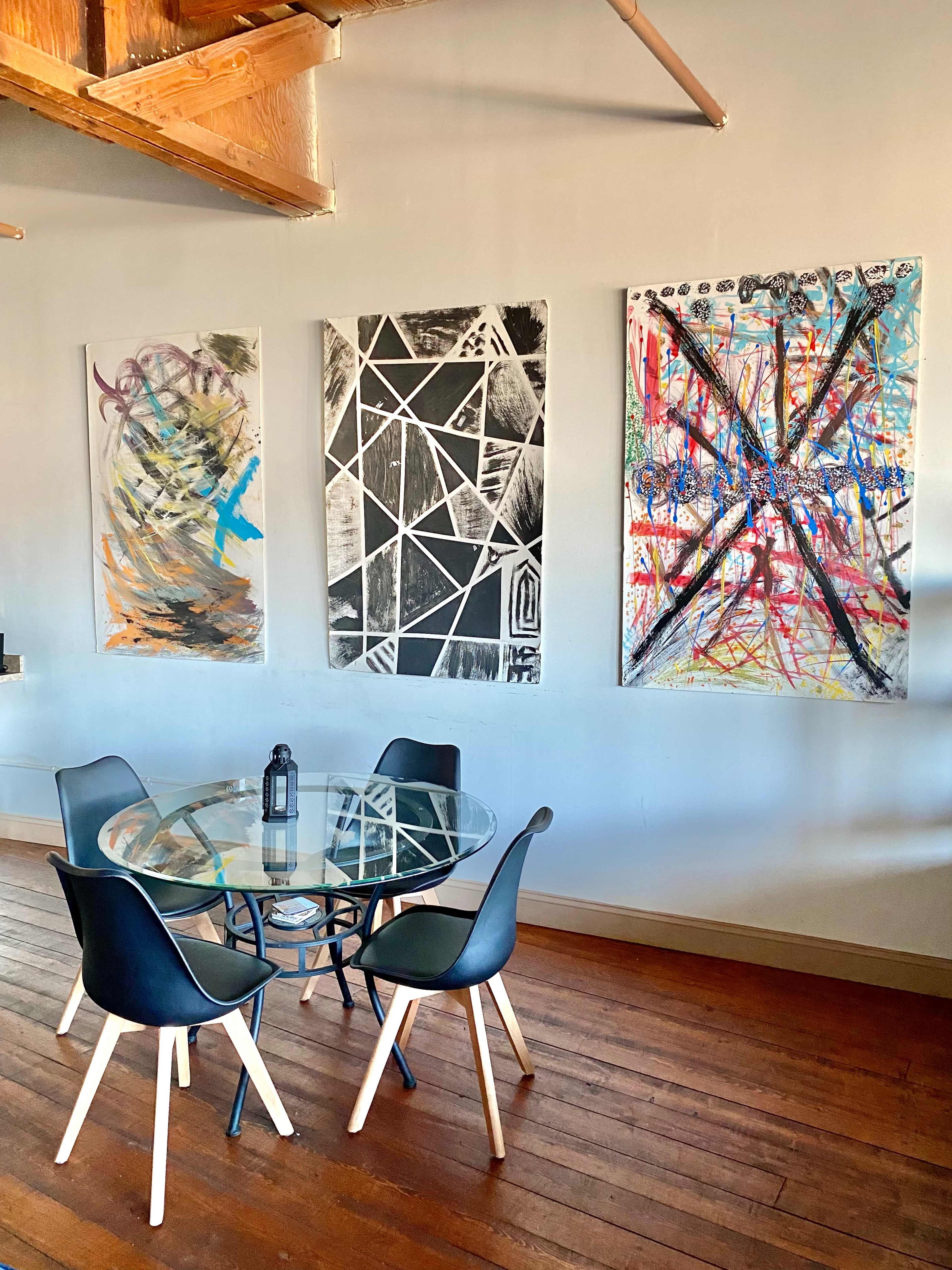 A glass dining table with black chairs is positioned on a wooden floor beneath three abstract paintings mounted on a gray wall.