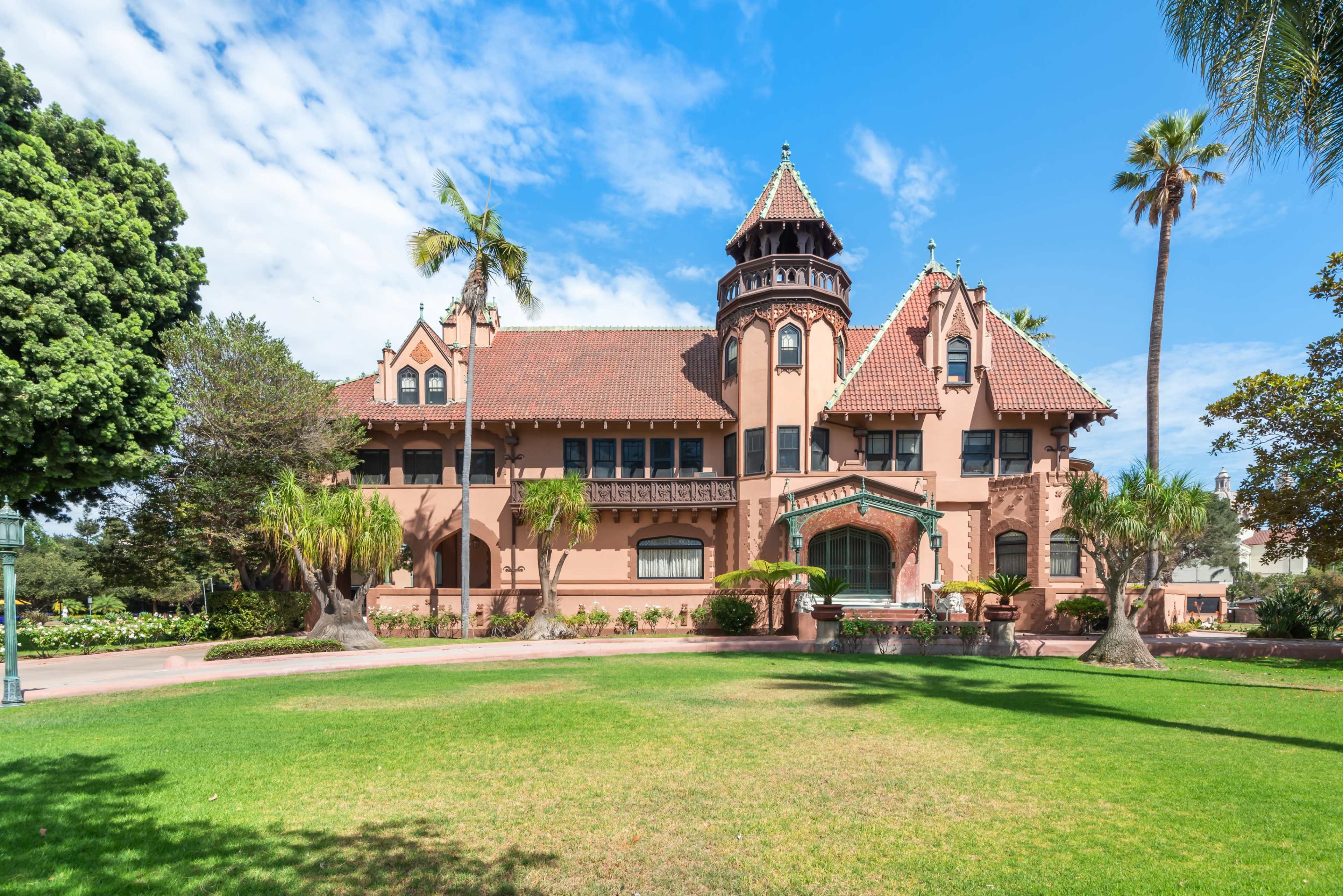 The image shows a large, historic mansion with a distinctive turret and ornate architectural details, surrounded by well-maintained lawns and palm trees.