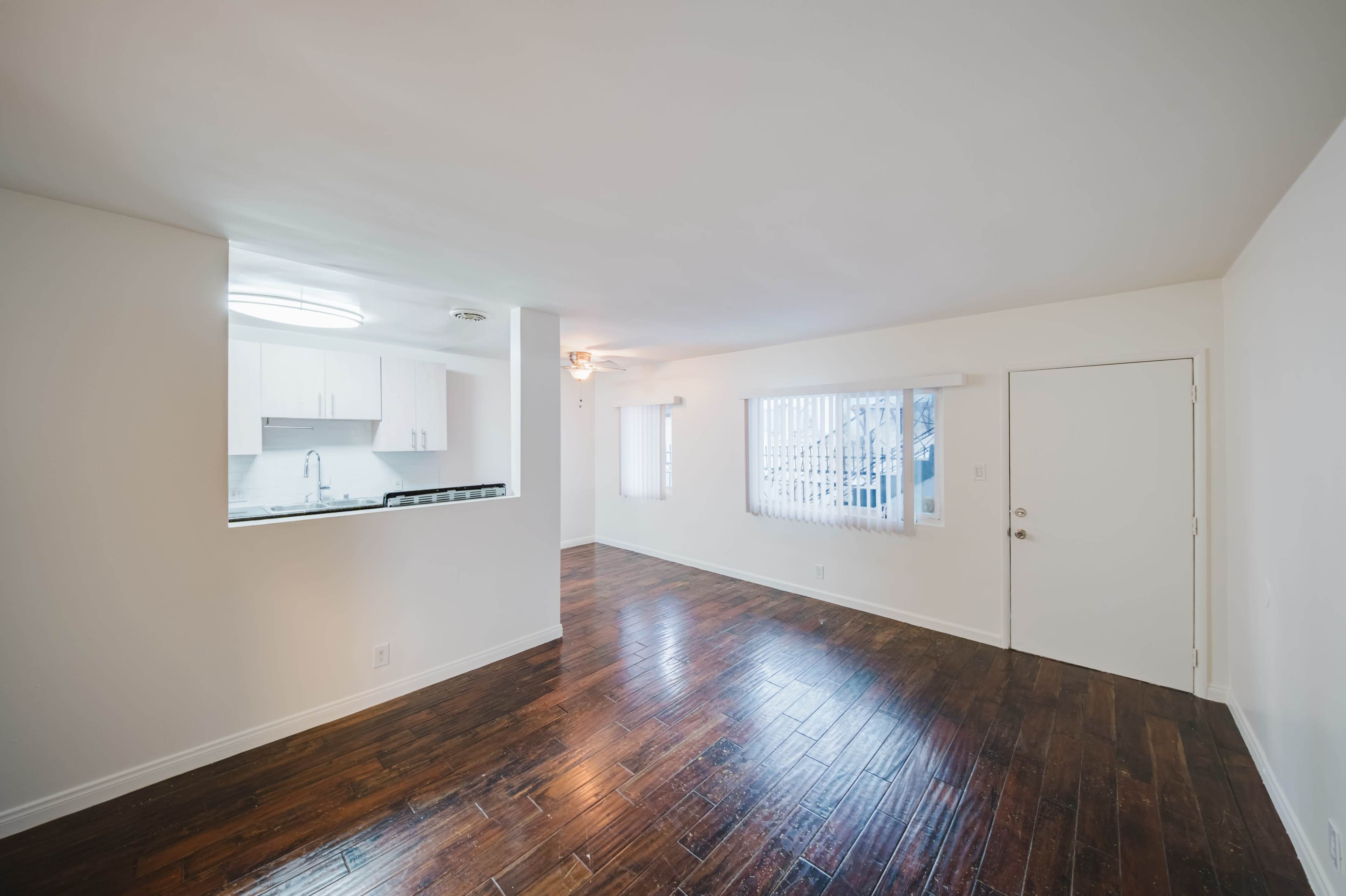 The image shows a bright, empty living space with hardwood floors, a kitchenette in the background, and a door leading outside.