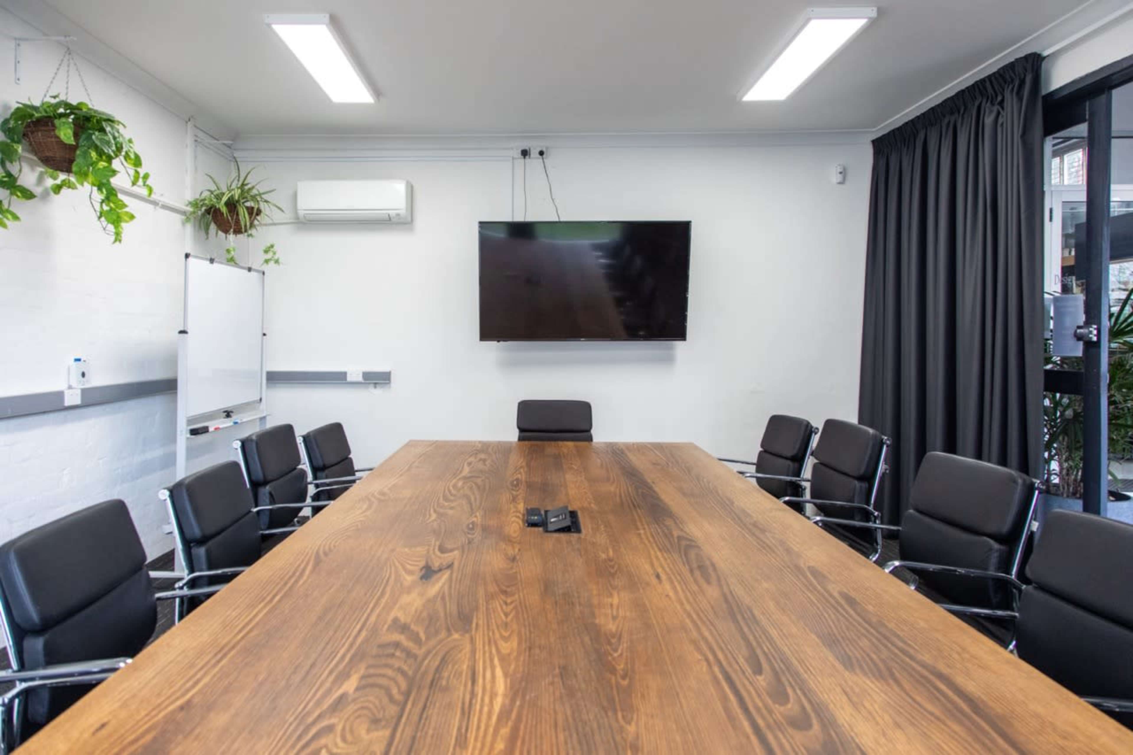 The image shows a modern conference room with a large wooden table, eight black chairs surrounding it, a wall-mounted television, and potted plants in the corners.