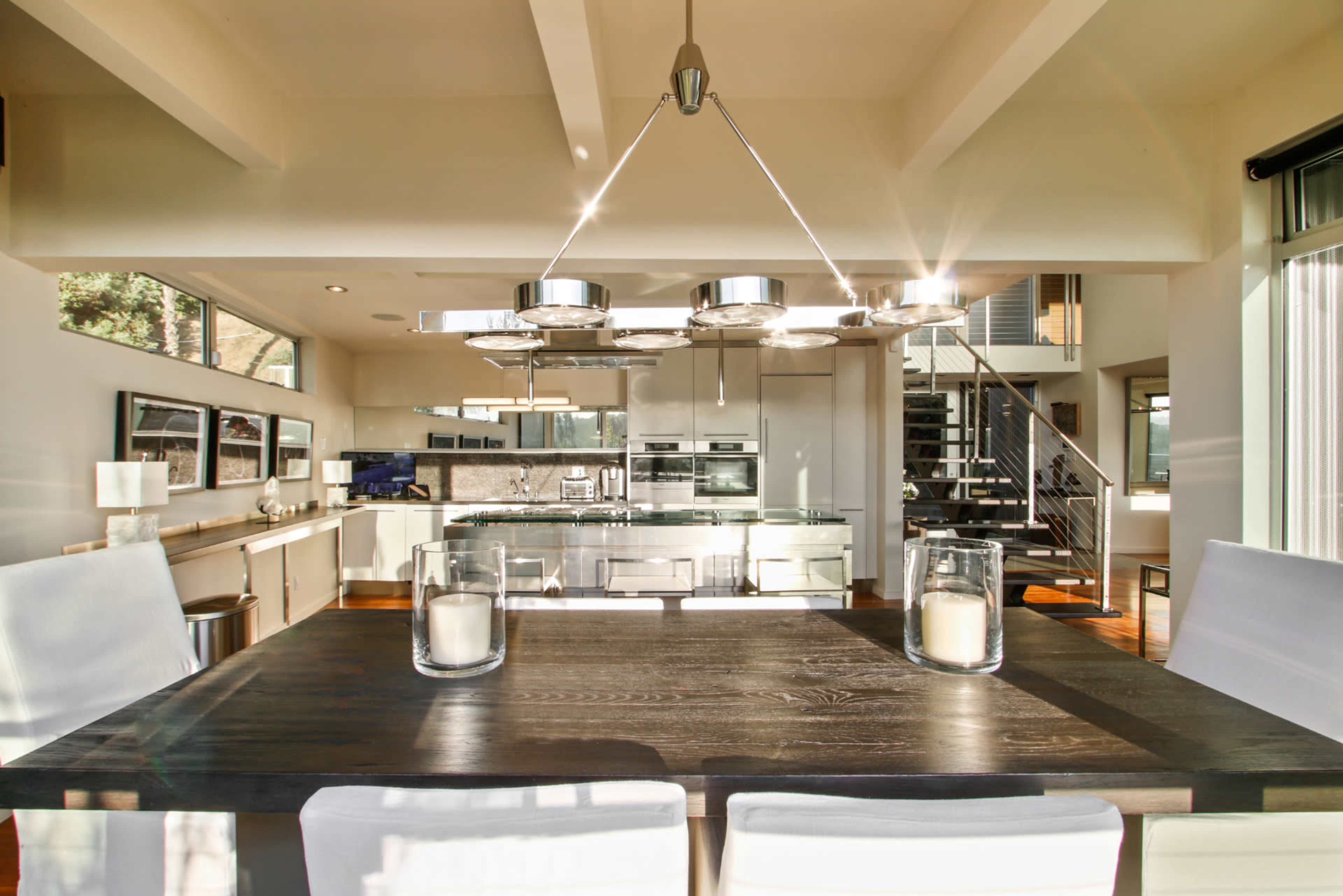 A modern kitchen and dining area features a large wooden table illuminated by overhead lighting, with sleek cabinetry and appliances in the background.